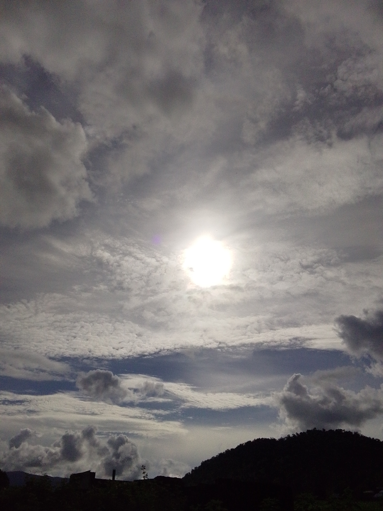 The image shows a vast, cloudy sky with the sun peeking through, creating a bright halo effect. The clouds vary in density and texture, with some appearing wispy and others more solid and dark. Below the sky, a silhouetted landscape of rolling hills and trees is visible against the bright sky, with a few dark, indistinct structures on the horizon. The overall scene suggests an outdoor environment, possibly in a rural or semi-rural setting. The crops provided do not offer any specific details about the location in Serti, Nigeria.