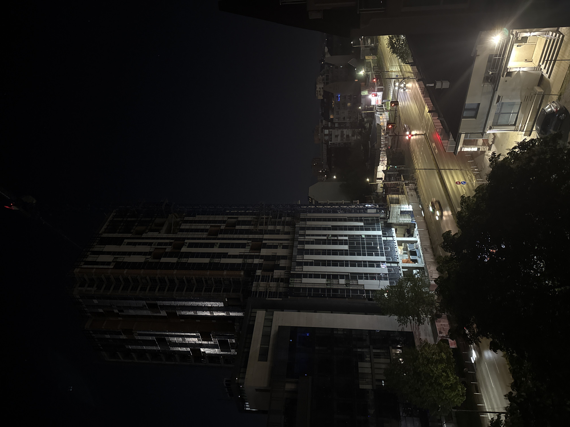 An outdoor night scene depicts a city street and construction site from an elevated vantage point, possibly a window. The left side shows a tall, unfinished building under construction, with visible scaffolding and partial cladding. The night sky appears dark.
To the right, a well-lit city street is visible, complete with traffic lights (showing red), streetlights, and blurred car lights indicating movement. Buildings line the street, some well-lit and others shadowed. Trees are also visible on the right side of the frame.