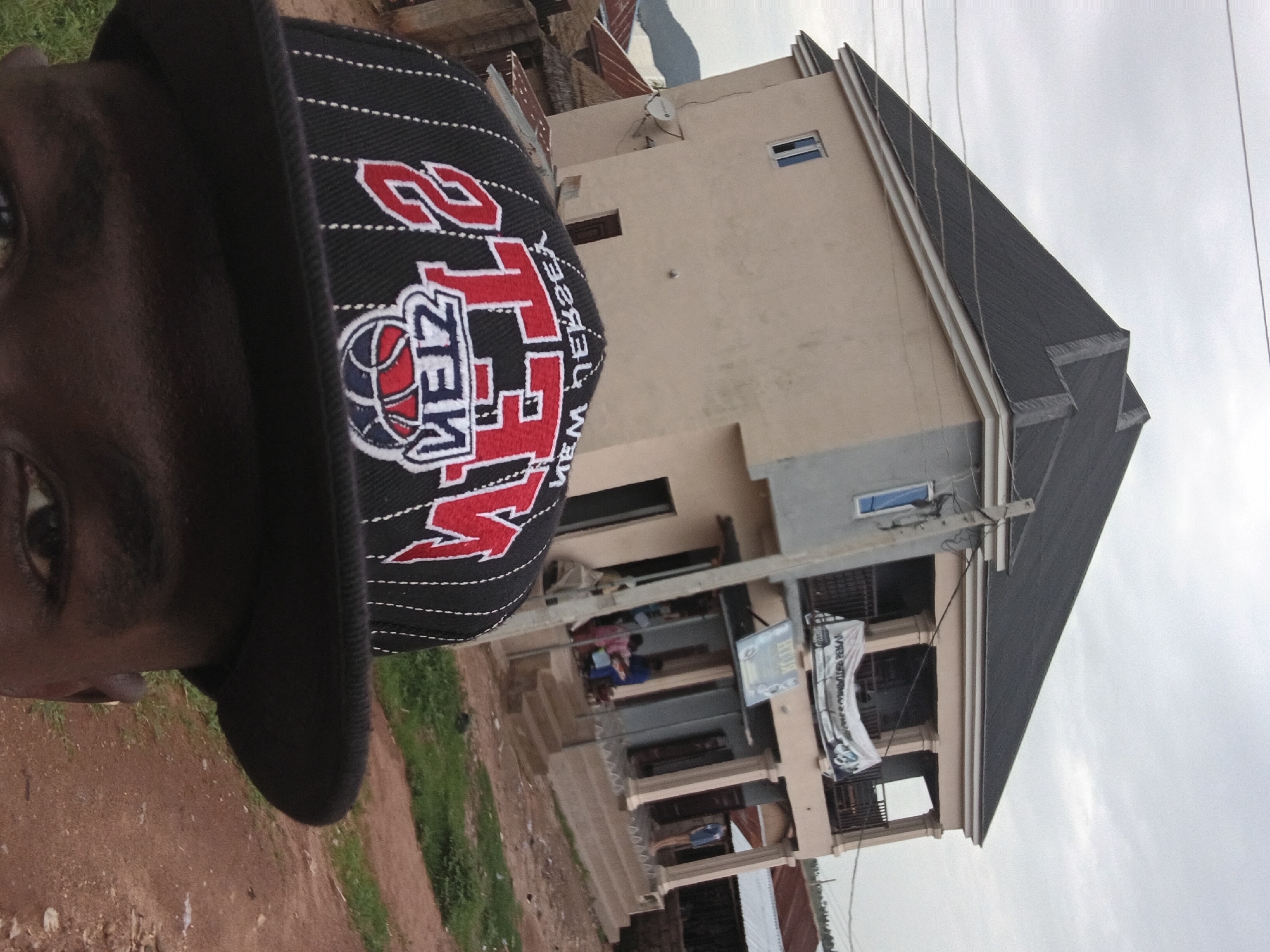 A selfie taken outdoors shows a person wearing a black baseball cap with red and white lettering. The cap has a graphic design featuring a basketball and the words "NJZIEM DEISEX". The person's face is partially visible in the foreground, with their eye and a portion of their cheek.
In the background, a multi-story building with a light-colored facade stands. The building has multiple windows, some of which are dark. There appears to be a balcony or covered area on the lower level, with people visible inside. A banner hangs from the building, displaying text, though it is difficult to read clearly. The building is situated on a dirt and grass-covered ground. A utility pole with wires is also visible.
The scene suggests a daytime setting, possibly in a semi-urban or rural area. The weather appears overcast. The image is oriented vertically, with the person's head at the top left and the building extending downwards and to the right.