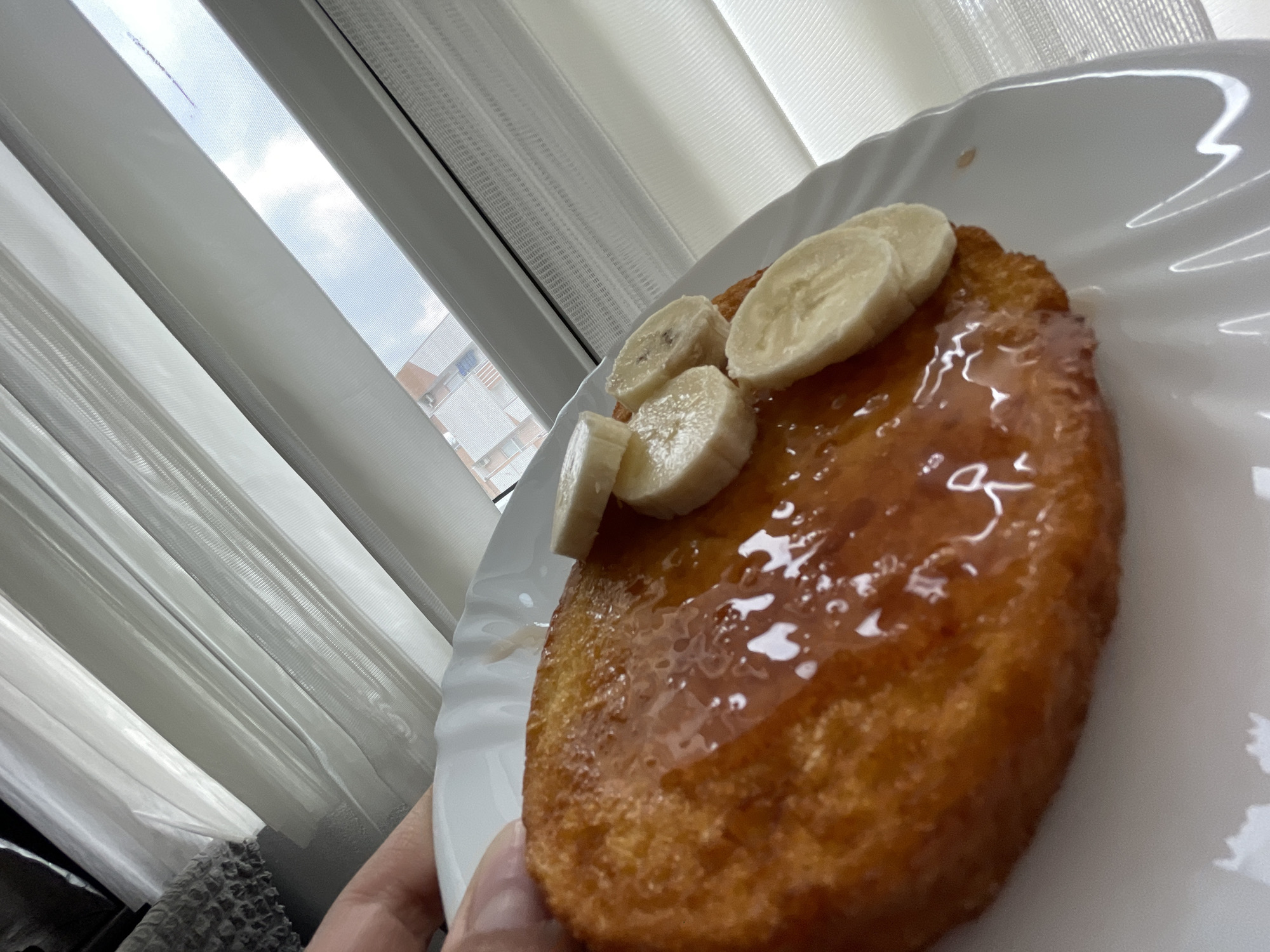 A close-up, angled shot shows a plate with what appears to be French toast topped with banana slices and syrup. The plate is white with a wavy edge. The French toast is golden brown and glistening with syrup. Several banana slices are arranged on top. The scene is lit by natural light coming through a window partially covered with white blinds. Outside the window, a building can be seen, hinting at an urban environment. The bottom left of the frame reveals part of a hand holding the plate and some fabric of a gray sweater. The location is indeterminable from the picture. Therefore, it is not possible to confirm that the picture was taken in Zlatograd, Bulgaria.