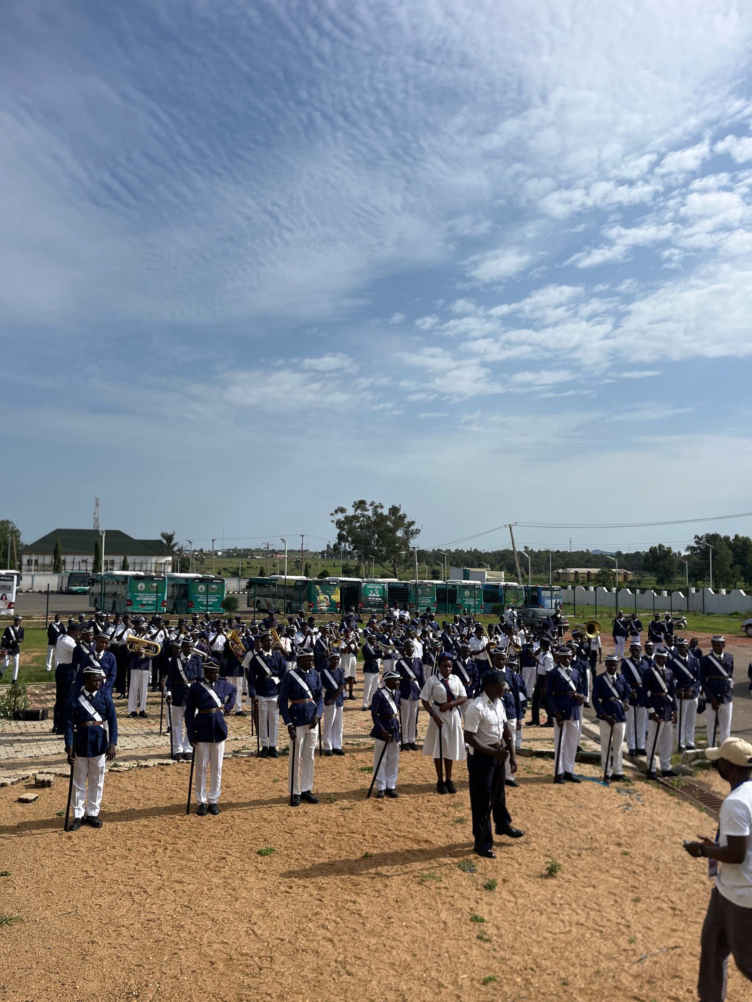 In Barikin Jirga Summa, Nigeria, a large marching band, composed of individuals in navy blue uniforms with white sashes and hats, stands at attention in a sandy outdoor area. Many members hold musical instruments, including trumpets and drums. Behind the band, a line of green buses with "40TH ANNIVERSARY" visible on their sides are parked, suggesting an event or celebration. In the background, there are buildings, trees, and a cloudy blue sky.