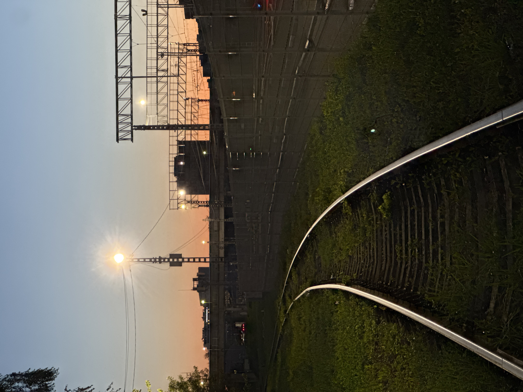 This image captures a railroad track curving to the left, surrounded by overgrown green grass. In the background, the sky displays a gradient of twilight colors, transitioning from a pale orange near the horizon to a clear blue above. A single bright street lamp illuminates the scene, casting a glow. Silhouetted against the twilight are various structures, including what appears to be a pedestrian overpass and industrial or large building complexes. Overhead, metal scaffolding structures, likely for the railway's electrical system, are visible. The overall impression is that of an industrial or urban fringe area at dusk.