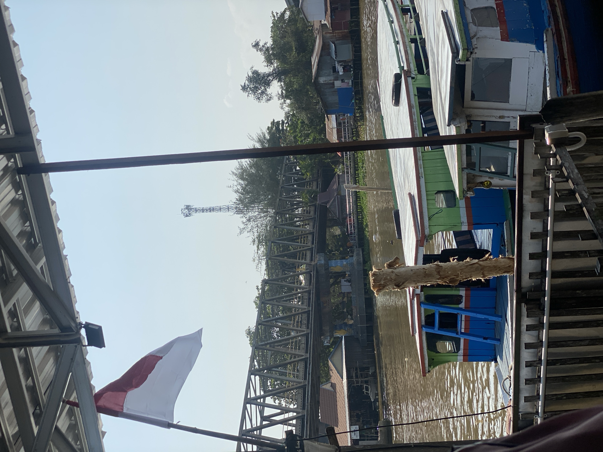 The image captures a view of the Martapura River in Banjarmasin, Indonesia, with several traditional wooden boats docked along the waterfront. The foreground features the roofs and upper structures of these boats, some painted in vibrant colors like green and blue. A rustic wooden ladder is visible on one of the boats. In the middle ground, a steel truss bridge spans the river, with a few buildings nestled along the riverbanks beneath it. Trees and vegetation line the river's edge, and a cell tower can be seen in the distance. The Indonesian flag is prominently displayed on a flagpole in the upper left portion of the image. The sky is clear and blue, suggesting a sunny day. The overall scene depicts a characteristic riverside environment of a Southeast Asian city.