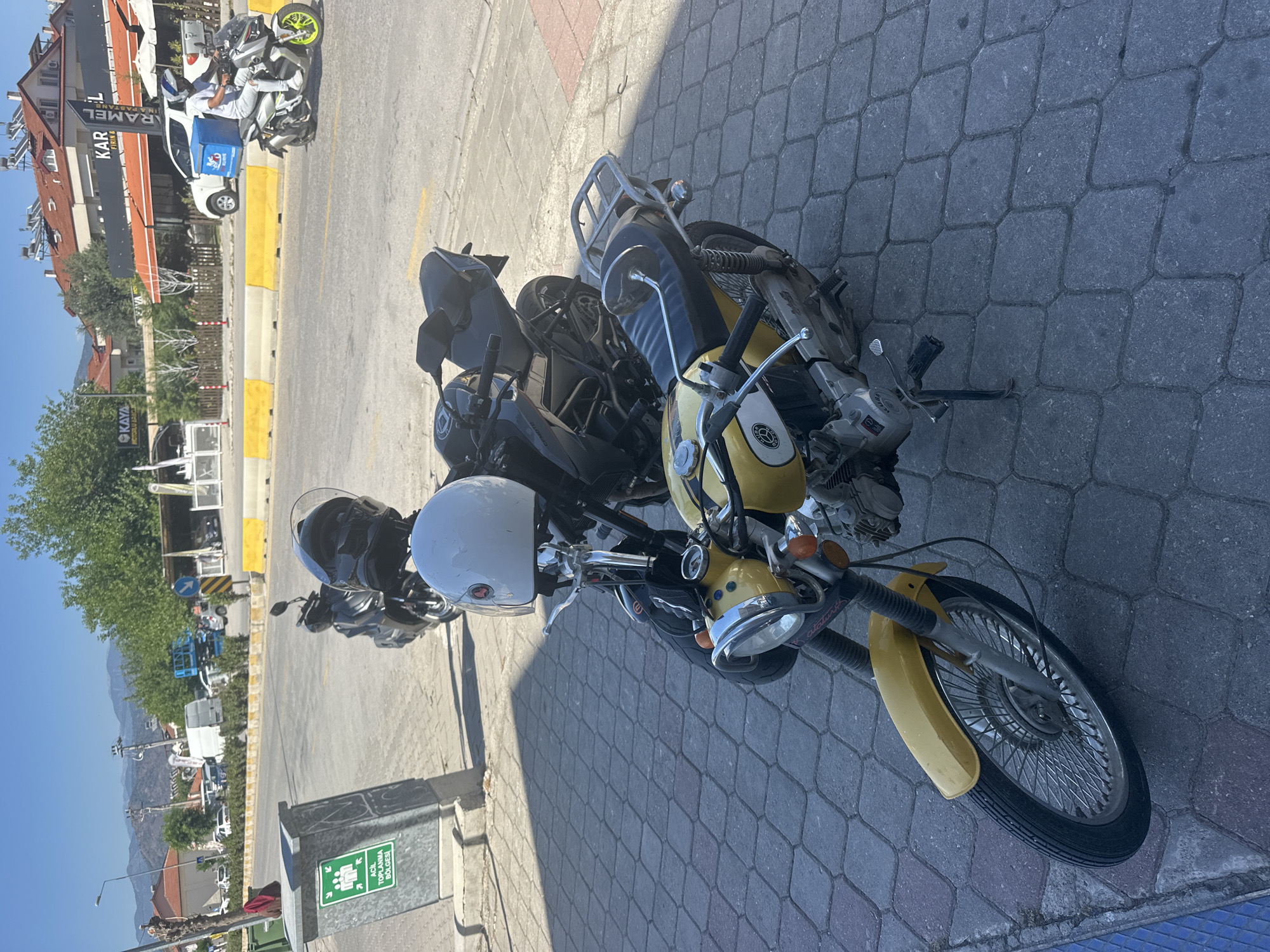 The image captures an outdoor scene on a sunny day, likely in Fethiye, Turkey, given the architecture, language, and mountainous backdrop. Several motorcycles are parked on a gray cobblestone sidewalk. In the foreground, a yellow classic-style motorcycle with prominent chrome headlights is visible, with a white helmet resting on its handlebars. Another dark-colored motorcycle is parked closely behind it.
To the left of the motorcycles, a street runs parallel to the sidewalk, separated by a yellow-painted curb divider. On the street, a white delivery van with a large blue box on its rear is parked, and next to it, a delivery scooter with a rider in a white outfit and helmet is seen. Further down the street, various cars and vehicles are visible.
In the midground, several buildings line the street. One prominent building features a sign that reads "KARAMEL," suggesting a business establishment. Another visible sign further down translates to "KAYA MOTEL & CAFE." Lush green trees are scattered along the street, providing shade.
In the background, majestic mountains rise under a clear, bright blue sky, indicating fair weather. The strong sunlight casts distinct shadows, suggesting it is mid-morning or afternoon. A gray waste bin on the sidewalk has a green sign with a white pictogram and the text "ACİL TOPLANMA BÖLGESİ," which translates to "Emergency Assembly Area." The overall scene depicts a busy, yet pleasant, street environment.