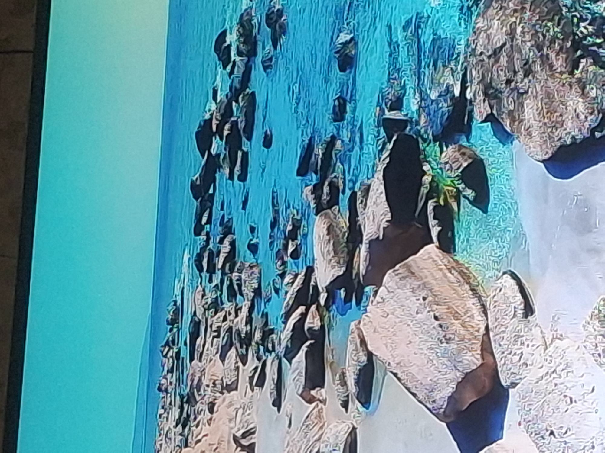 The image shows a rocky coastline with clear turquoise water. Large, weathered stones and boulders are scattered along the shoreline and in the shallow water. The sand appears light in color. The horizon shows a faint line of land under a bright, clear blue sky. There are no people or man-made structures visible in the scene. The image does not depict the city of Izmail, Ukraine.