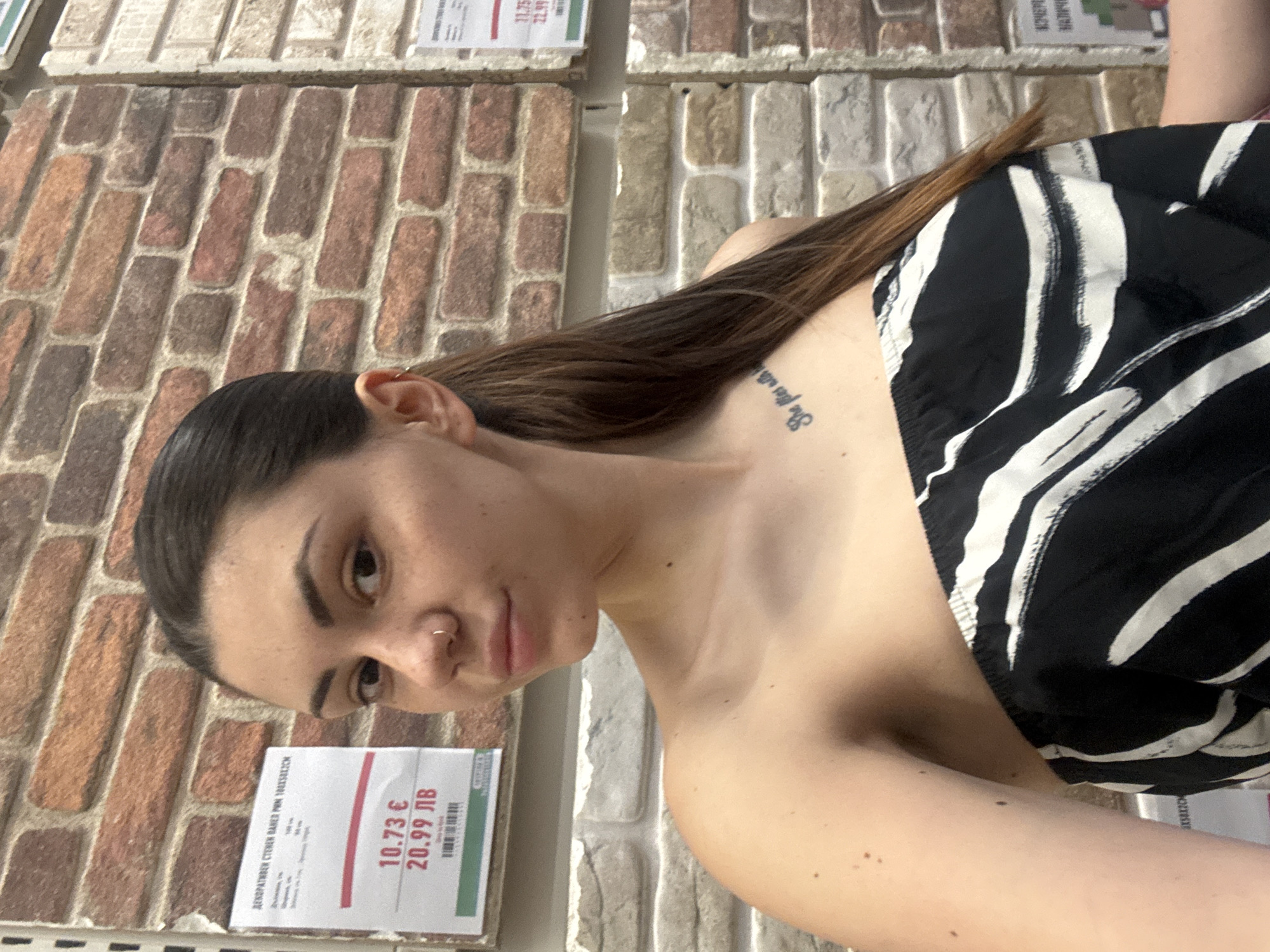 A young adult female is depicted in a medium close-up, posing in what appears to be a retail store, likely located in Sofia, Bulgaria. The background consists of various sample panels exhibiting a brick-patterned design, consistent with a home improvement or building materials establishment.
The female subject has dark brown hair, pulled back into a ponytail with lighter brown undertones visible. She has dark eyebrows and a small silver hoop nose ring in her left nostril. Her skin displays visible freckles, particularly on her face and left shoulder. She is wearing a strapless or off-shoulder top with a black and white abstract stripe or zebra print pattern. A script tattoo is visible on her left clavicle, partially obscured but appearing to read "She flies with...". A small silver hoop earring is present in her left ear. Her gaze is directed towards the camera, suggesting a self-portrait.
To her left, a white sign with black and red text displays product information, including "ДЕКОРАТИВЕН СТЕНЕН ПАНЕЛ" (Decorative Wall Panel) and prices: "10.73 €" and "20.99 ЛВ". The "ЛВ" currency indicator confirms the location is in Bulgaria. A similar, partially visible sign is on the right side of the frame. The lighting is consistent with indoor retail environments.