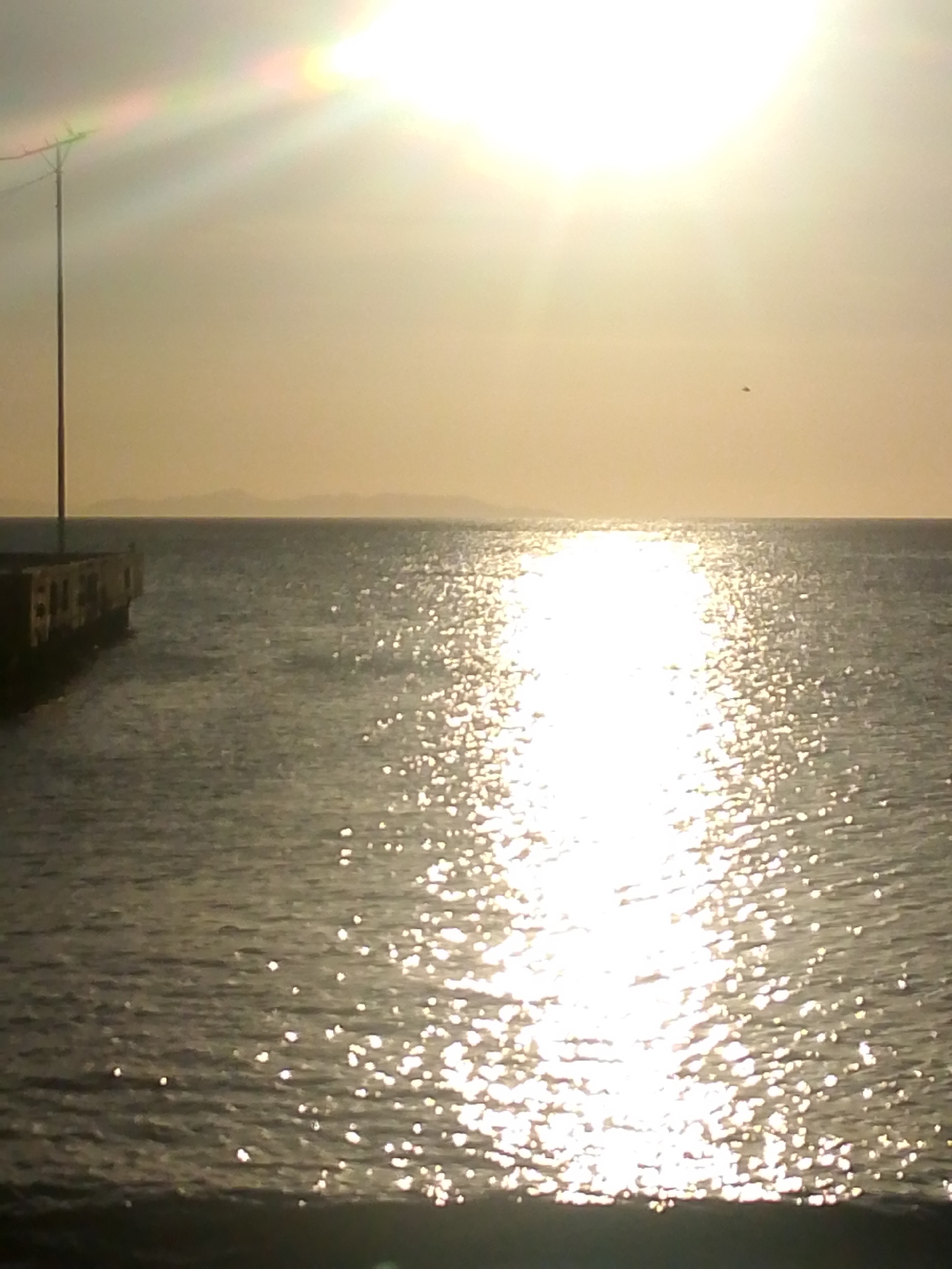The image depicts a coastal scene in Juan Griego, Venezuela, under conditions of intense backlighting, likely during sunrise or sunset. The sun is prominently positioned in the upper-right portion of the frame, casting a strong, glinting reflection path across the water that extends from the horizon towards the bottom-center. Lens flare is visible in the upper-left sky, which appears hazy and yellowish-orange. A small, dark, indistinct object is present in the upper-right sky. A distant landmass is faintly silhouetted on the horizon. On the far left, a tall, slender pole, possibly a utility or antenna mast, extends vertically into the sky, featuring a cross-arm structure at its apex. Below this pole, a dark, concrete structure, consistent with a pier or seawall, extends into the water from the left edge of the image. The water's surface exhibits ripples and waves, with areas not directly in the sun's reflection appearing darker. No individuals are visible, and no specific actions or interactions are discernible.
