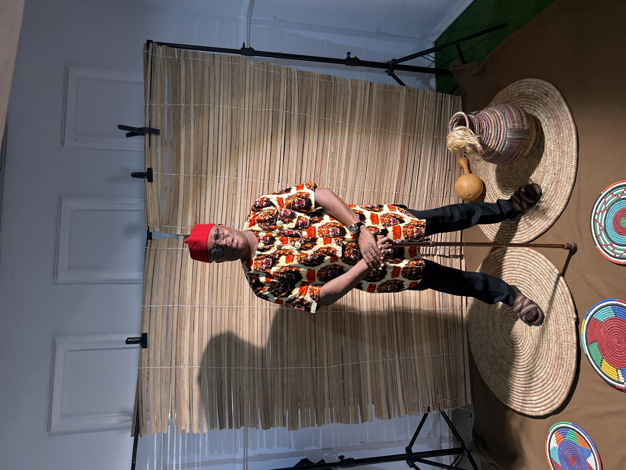 A man in a red cap and patterned shirt stands in front of a bamboo screen. He is wearing glasses, a watch, and sandals. The scene includes various woven items, such as baskets and mats, placed on a brown surface. In the background, there is a wall with architectural details and a patch of artificial grass above the bamboo screen. The location appears to be a studio setting and it is not possible to confirm that it is in Jos, Nigeria.