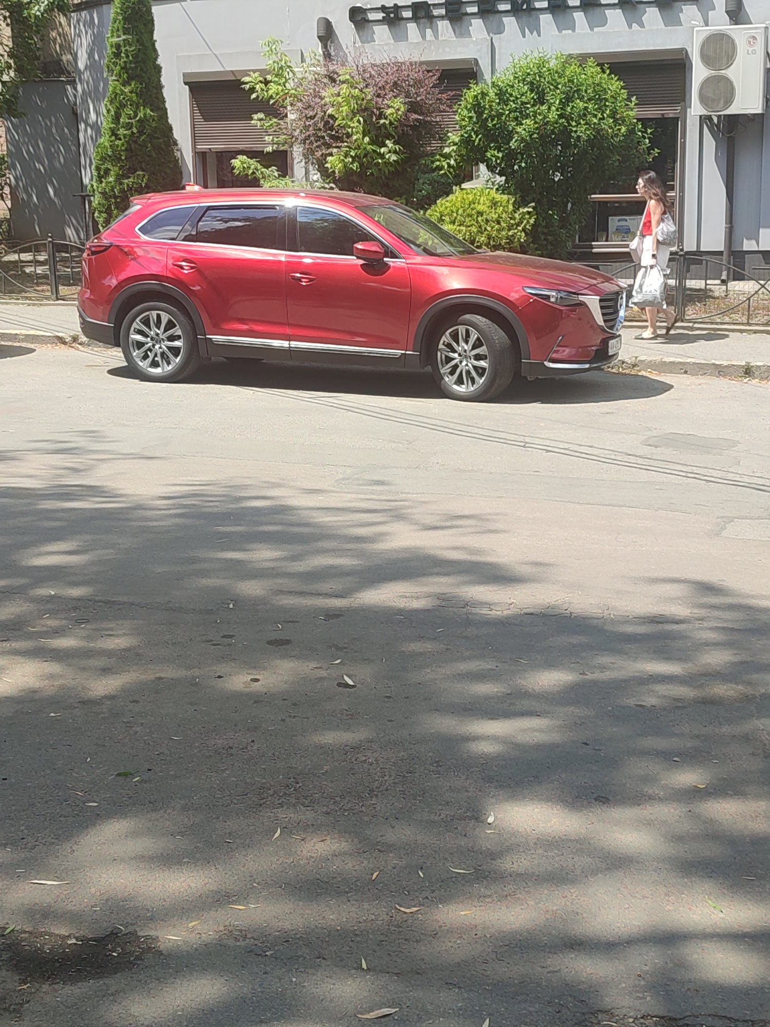 A red SUV is parked on the side of a street in Chernivtsi, Ukraine. A woman is walking on the sidewalk in front of a building with large windows and an air conditioning unit. The street is paved and has some cracks and patches. Tree branches cast shadows on the road. The weather appears to be sunny.