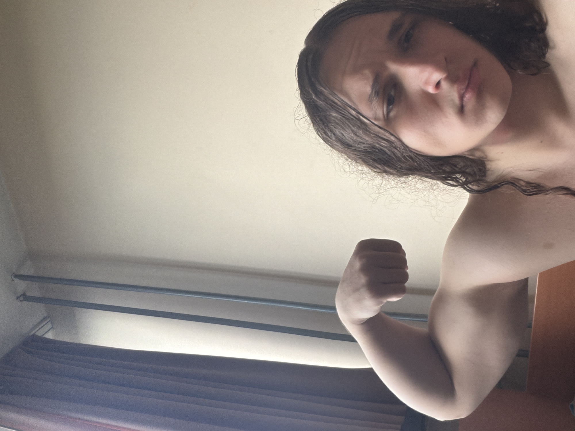 A low-angle selfie shows a person with long, wet brown hair flexing their right bicep. The person has fair skin. Behind the person, there is a beige-colored wall and what appear to be metal rods for hanging curtains. A purple-colored curtain is visible at the bottom of the frame. There is no verifiable evidence within this image to pinpoint a specific location in Plovdiv, Bulgaria.