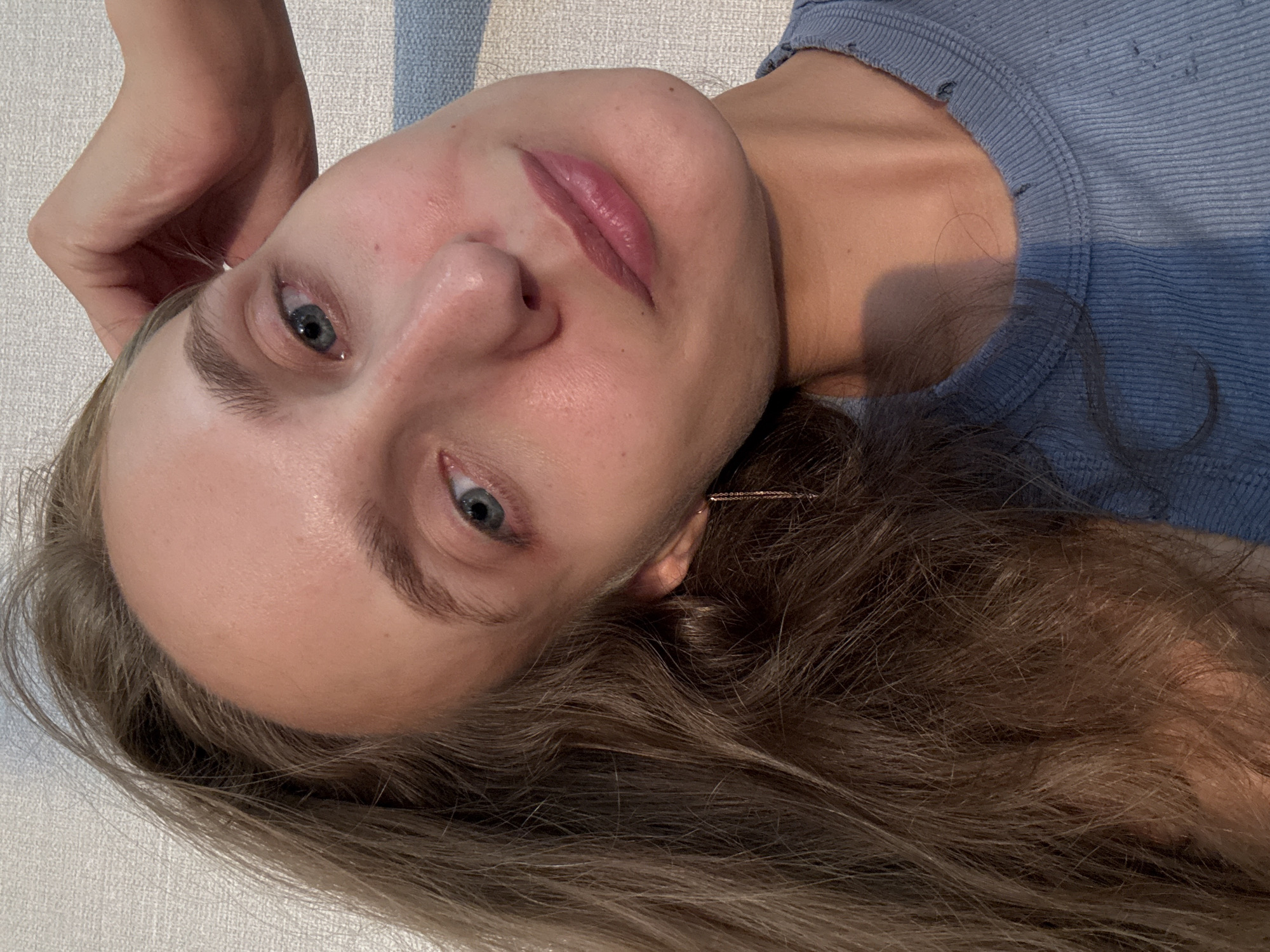 The image is a close-up, low-angle selfie of a young Caucasian woman with long, wavy brown hair and light blue eyes. She is wearing a blue ribbed tank top. Her left hand is resting on the back of her head, and she is looking directly at the camera with a neutral expression. The background appears to be a textured wall with soft lighting. There are no discernible elements within the image that would allow for the identification of a specific location in the city of Подшивалово, Russia.