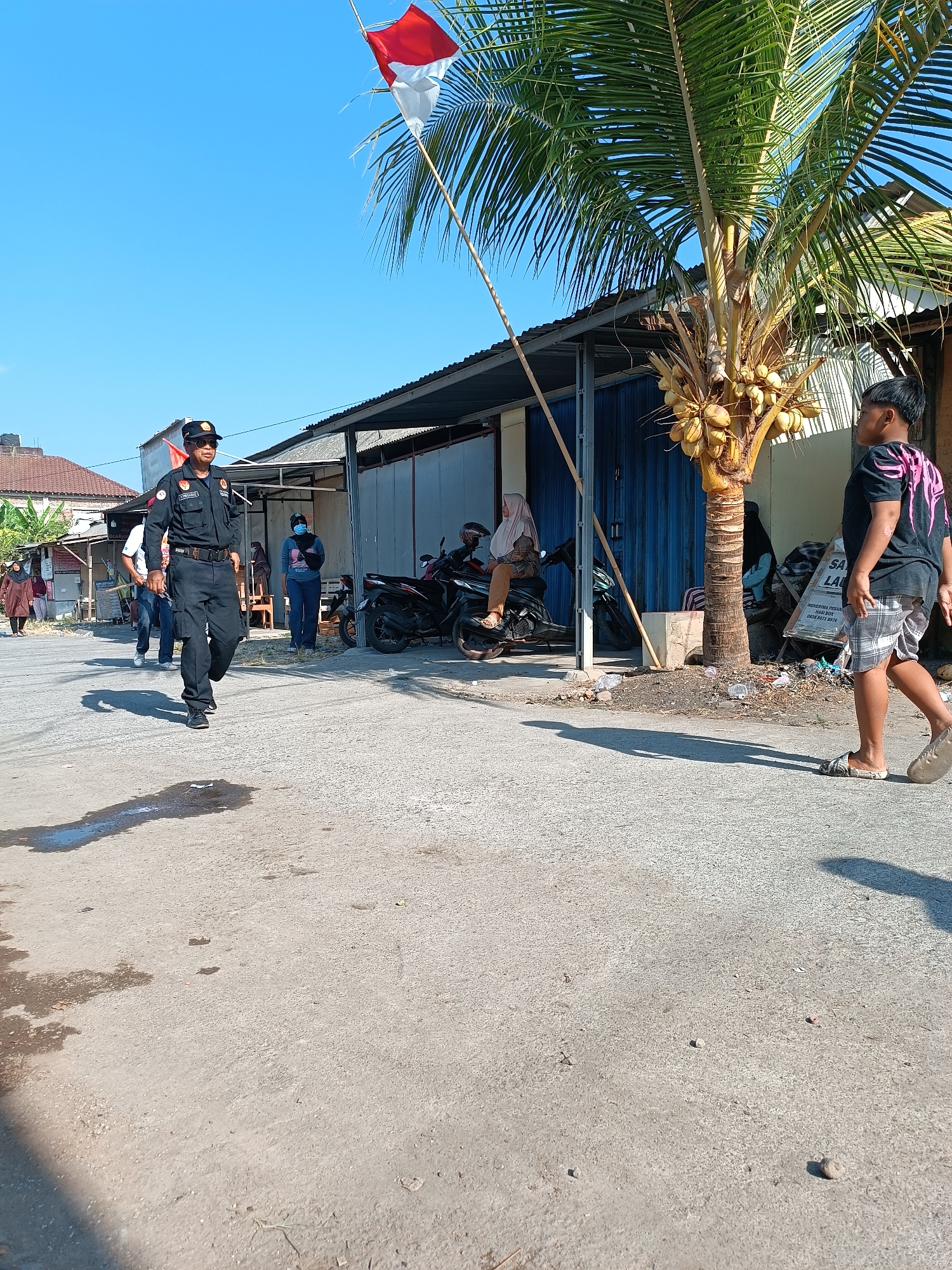 In Imogiri, Indonesia, a man in a black uniform, wearing a cap and sunglasses, walks towards the viewer on a paved road. He has a serious expression and is holding what appears to be a red flag in his left hand. Behind him, several other individuals are visible, including a woman wearing a hijab and a blue shirt, standing near motorcycles. To the right, a young boy in a black t-shirt and plaid shorts walks away from the camera, his back to the viewer. A coconut tree with numerous yellow coconuts stands prominently in the foreground, casting a shadow on the road. The scene depicts a street environment with buildings lining the background and a clear blue sky overhead.