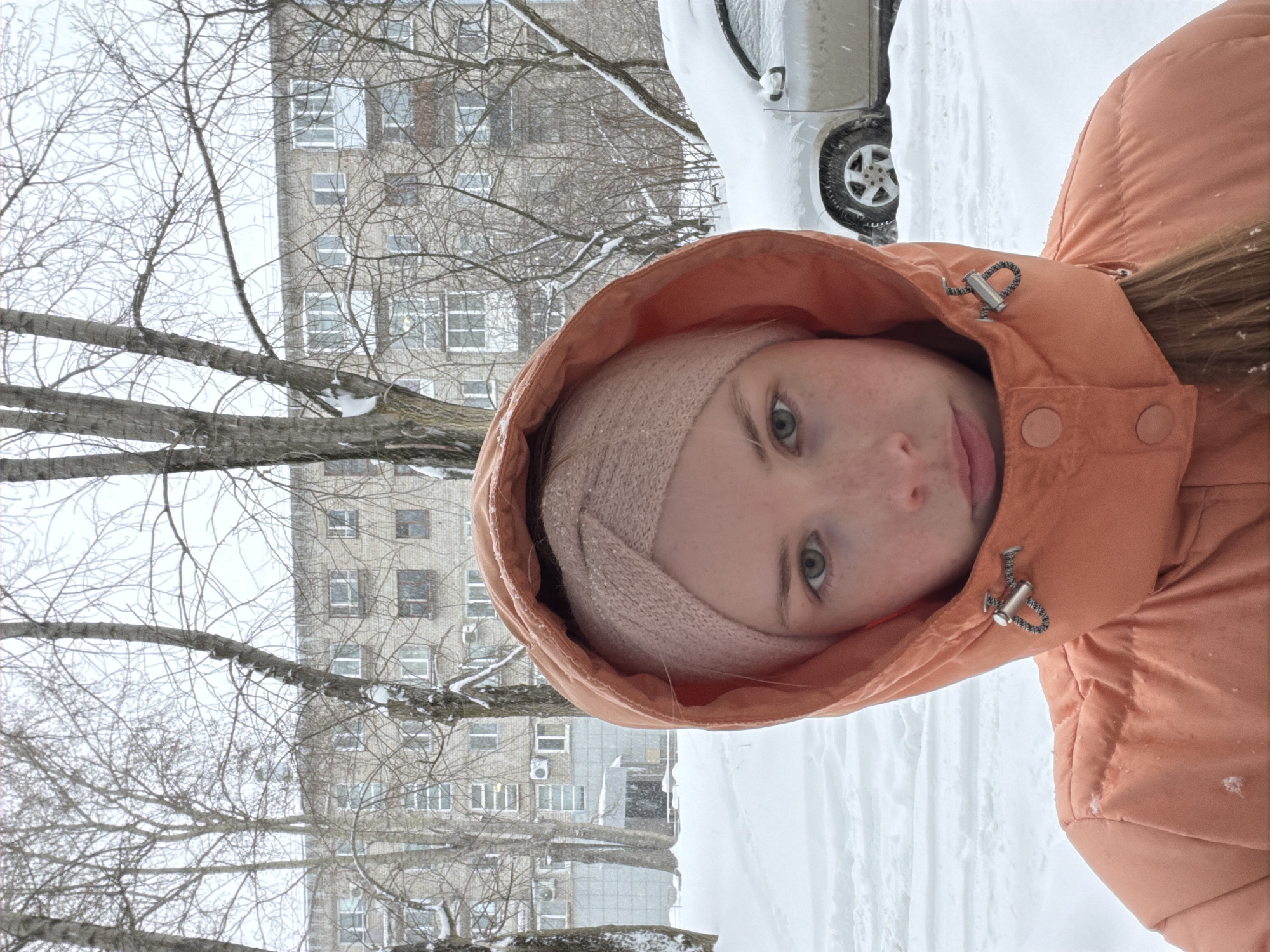 This media file captures a close-up portrait of a young woman outdoors in a snowy, urban setting in Berezniki, Russia. The image is oriented sideways and, if rotated upright, shows the woman on the right side, partially obscured by her peach-colored hooded winter jacket. She wears a light pink or peach knit beanie under her hood, and only her right eye, nose, and mouth are visible as she looks towards the camera with a neutral expression.
The background reveals a winter landscape dominated by snow. The ground is heavily covered in white snow, indicating cold temperatures and recent snowfall. A silver or light-colored car, also partially covered in snow, is visible behind her to the upper right. Leafless trees with intricate bare branches fill the middle ground, some branches dusted with snow, obscuring parts of a multi-story residential building in the distance. The building is constructed from light-colored bricks or panels and features numerous rectangular windows, with some air conditioning units attached to its exterior. The sky appears bright and overcast, consistent with daytime during winter weather. No discernible text is visible in the image. The scene conveys a typical cold, snowy day in a residential area.