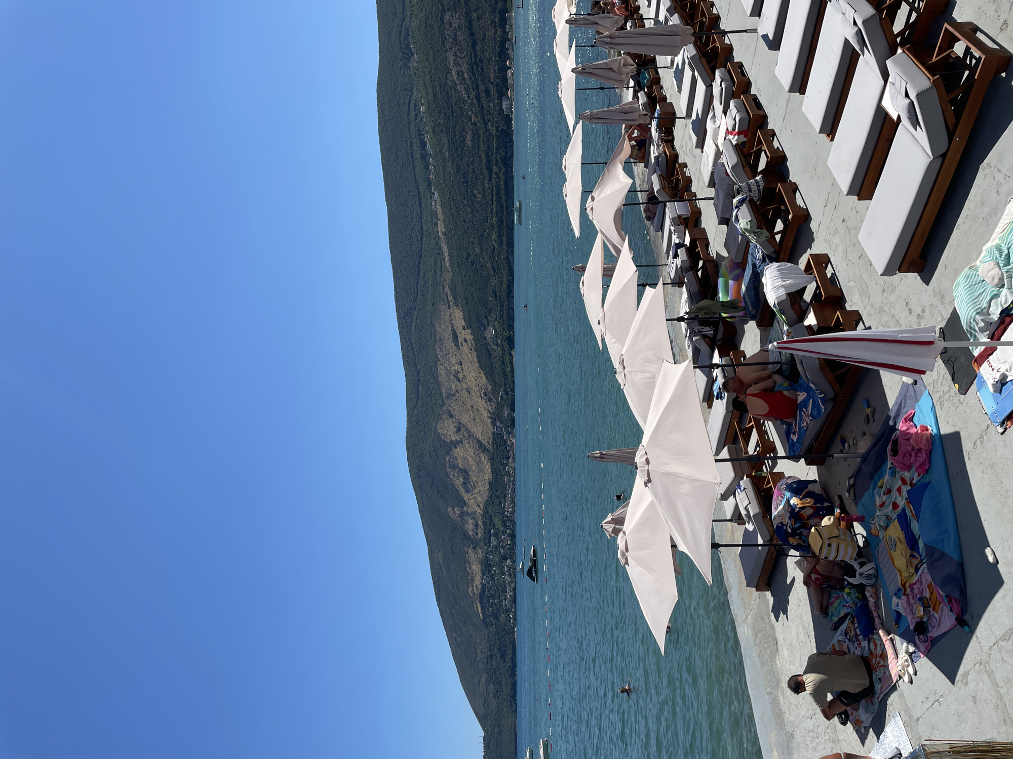 The image shows a beach scene in Igalo, Montenegro, on a clear, sunny day. In the foreground, numerous lounge chairs with grey cushions are arranged in rows on a concrete surface. Many of these chairs are unoccupied, while some are occupied by people. Several beach umbrellas, mostly light-colored and open, provide shade. In the immediate foreground, towels, bags, and a cooler are spread out on the ground, with a person sitting on a towel, facing away from the camera. Further back, people are seen lounging on chairs, and some are in the water. The sea is a vibrant blue, with a few boats visible in the distance. A long, tree-covered mountain range forms the backdrop, with some structures visible on its slopes and a town nestled along the coast. The overall atmosphere is one of a relaxed beach resort.