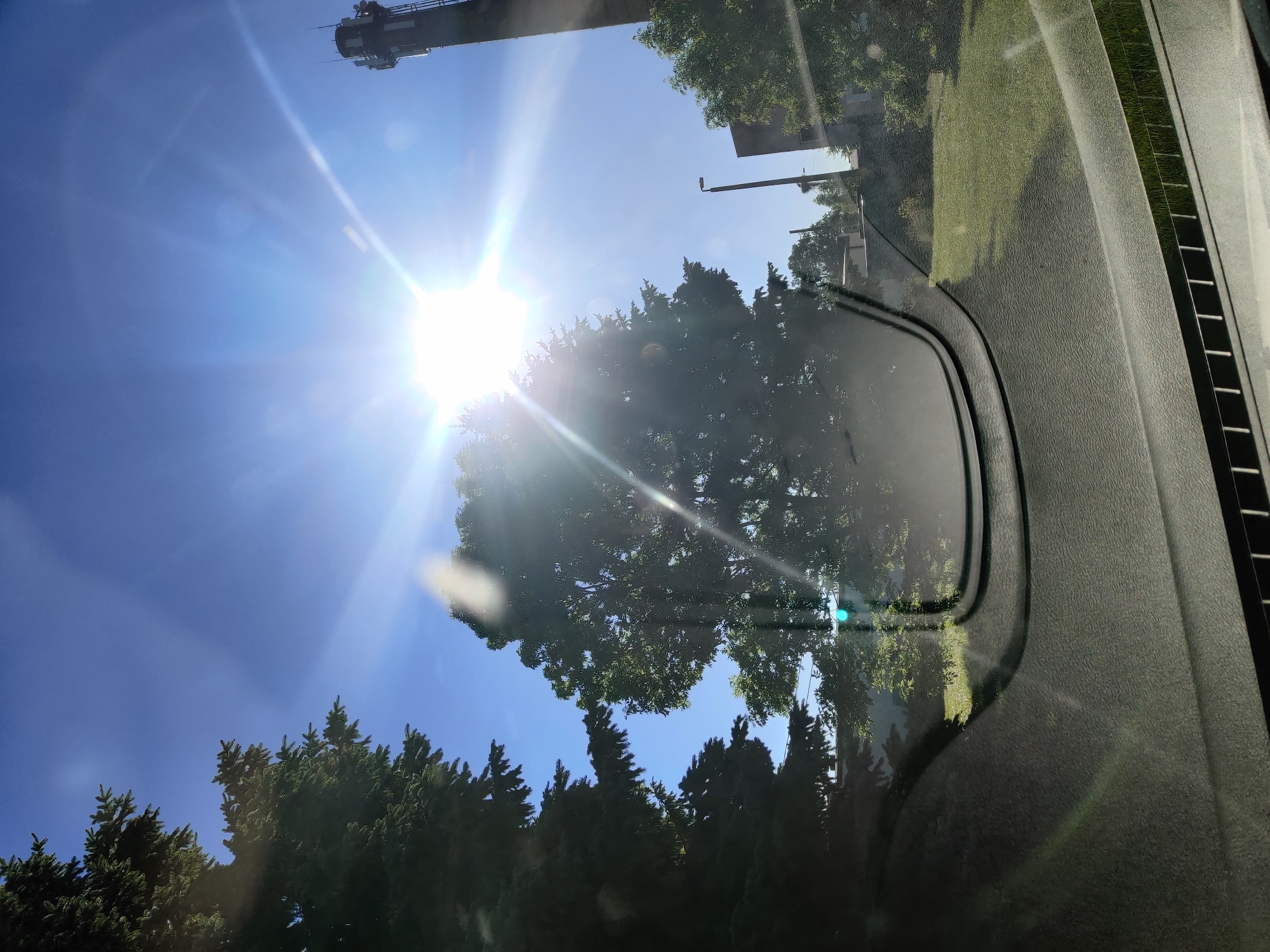 The image, captured from inside a vehicle looking out through the windshield, shows a bright sunny day. The sun is intensely bright, creating lens flare and glare across the image. On the left, a large evergreen tree dominates the foreground. In the center, another large deciduous tree is partially obscured by the sun's glare. To the right, a tall industrial tower, likely a communication or industrial chimney, is visible against the clear blue sky. A modern street lamp and some low buildings are also present in the background. The view suggests an outdoor setting, possibly in an industrial or suburban area of Saue, Estonia, given the presence of the tower and the general landscape. The lower portion of the image shows the interior of the car's dashboard and windshield, with reflections and glare. There are no people visible in the image.