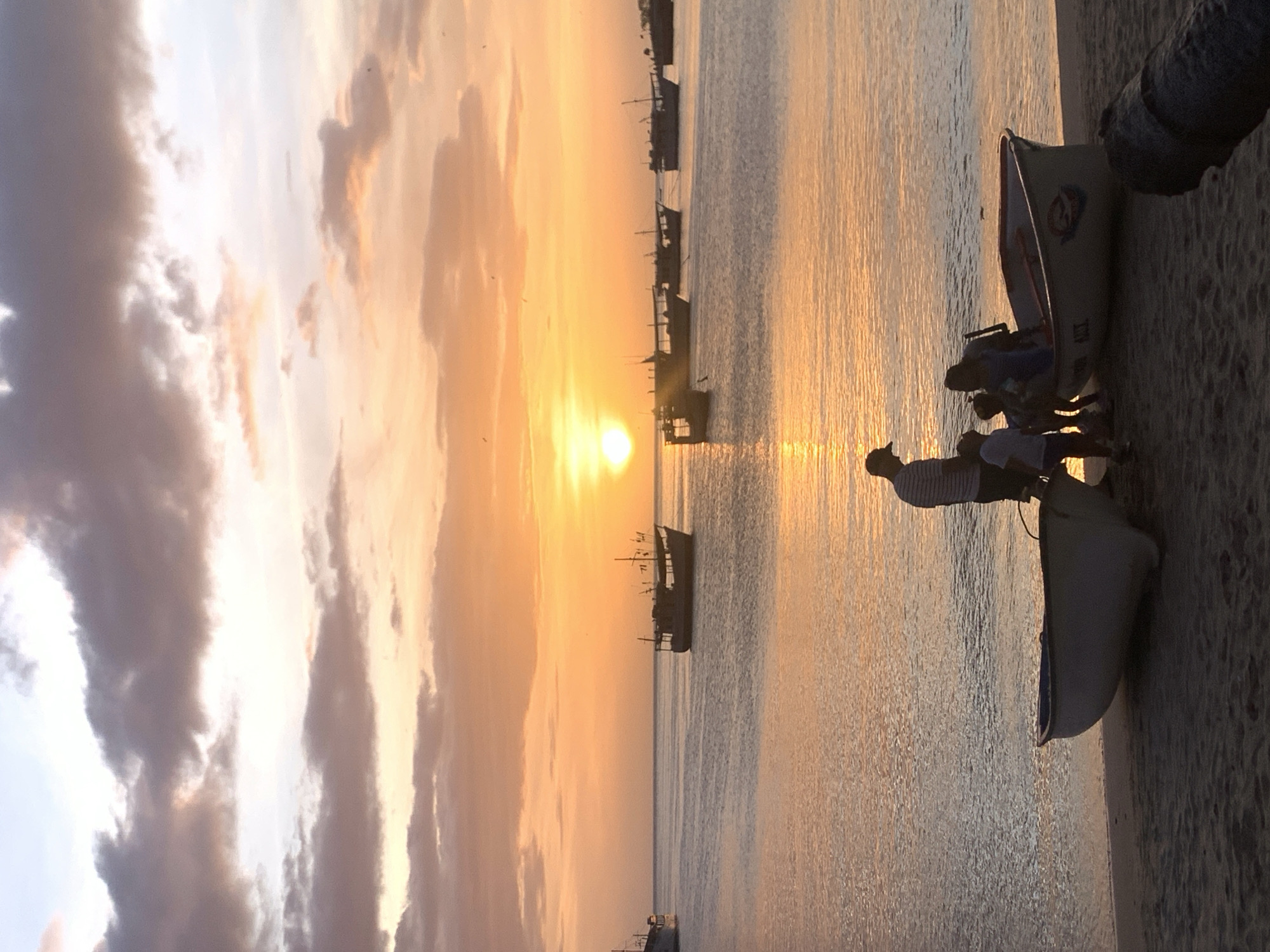 A family is gathered around a small boat on the sandy shore of Juan Griego, Venezuela, during sunset. The sky is ablaze with hues of orange, yellow, and pink, with dramatic clouds silhouetted against the vibrant sunset. The ocean reflects the colorful sky, creating a shimmering surface. Several fishing boats are anchored in the distance, adding to the picturesque scene. The family, composed of two adults and three children, appears to be preparing to embark or disembark from the boat. One adult is standing, facing the water, while the other adult and children are within or near the boat. The atmosphere is serene and tranquil, with the setting sun casting a warm glow over the entire landscape.