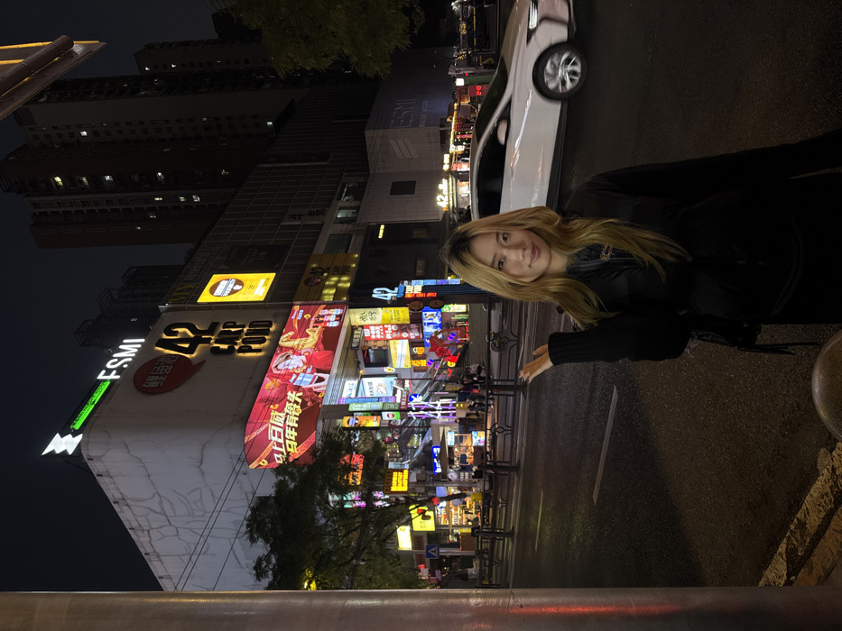 A young Asian female with shoulder-length blonde hair and a black padded jacket stands on an asphalt street in Liangjiang, China, at night. She faces forward, smiling subtly, with her right hand raised to her side. To her immediate left is a large grey cylindrical pole. The street behind her features white lane markings and black metal bollards separating the road from the sidewalk. A blurred white vehicle is in motion on the road to her right.
In the background, a large, multi-story building dominates the scene, featuring a light-colored facade. Illuminated signs on this building display "FSMI" with a green conical icon on the roofline, and "42 CARP POND" with a red fish logo below it. A large digital billboard underneath "42 CARP POND" shows a colorful advertisement with figures and Chinese characters. The building's lower levels and an entrance area are brightly lit by numerous colorful neon signs and smaller displays. Beyond this, taller, darker high-rise buildings with scattered illuminated windows are visible against the dark night sky. Other pedestrians are faintly visible in the brightly lit areas of the building's entrance.