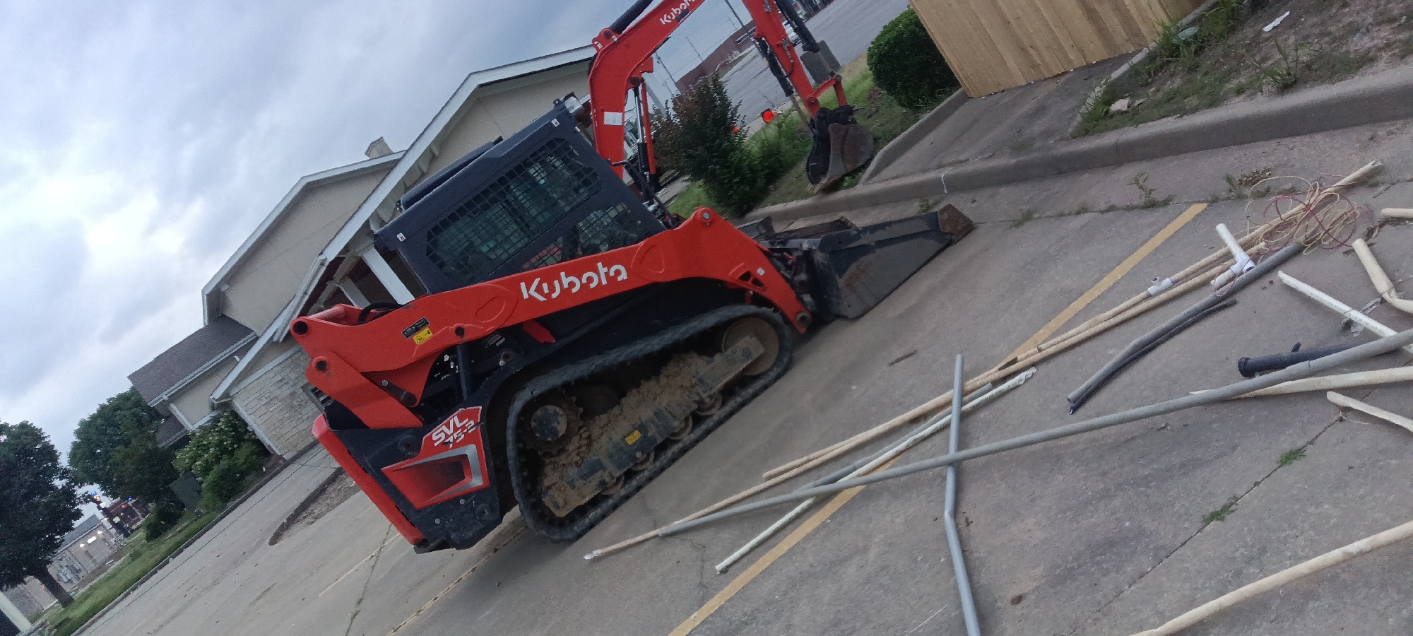 A Kubota SVL75-2 compact track loader is on a concrete surface next to a curb. Several cut or broken poles and rods are on the concrete surface around the machinery. In the background are buildings, greenery, and a wooden fence. The scene is potentially in or near Van Buren, United States of America.