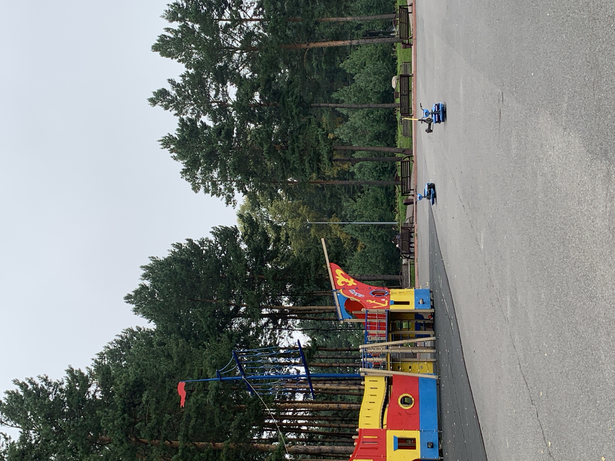 The image depicts an outdoor playground scene with a pirate ship-themed play structure in the foreground. The structure is brightly colored in red, yellow, and blue, with a blue mast and red flag. Behind the playground, tall pine trees form a dense forest, characteristic of the natural landscape in Prokopyevsk, Russia. Several wooden benches are arranged in a row in front of the forest, and a few people are seated on them. Two blue electric scooters are present on the asphalted area in front of the benches, with one appearing to be in motion. The overall environment suggests a park or recreational area.