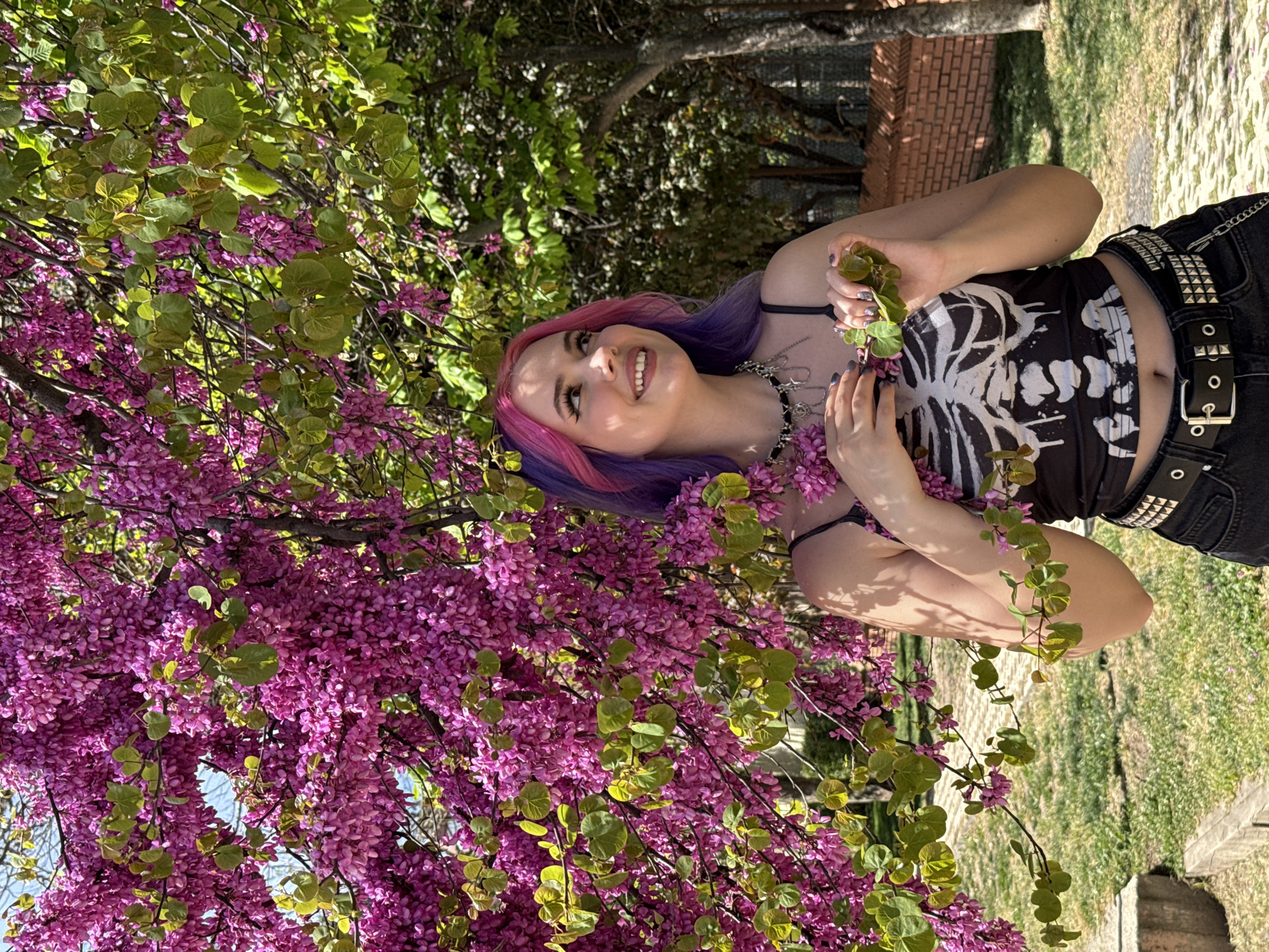 A young woman with pink and purple hair smiles in front of a blooming tree with pink flowers. She wears a black crop top with a white skeletal design, black studded jeans, and a silver chain necklace. The location appears to be a park in Madrid, Spain, with a brick wall in the background and green grass on the ground. The tree's flowers are the focal point, obscuring part of the woman's face and body.