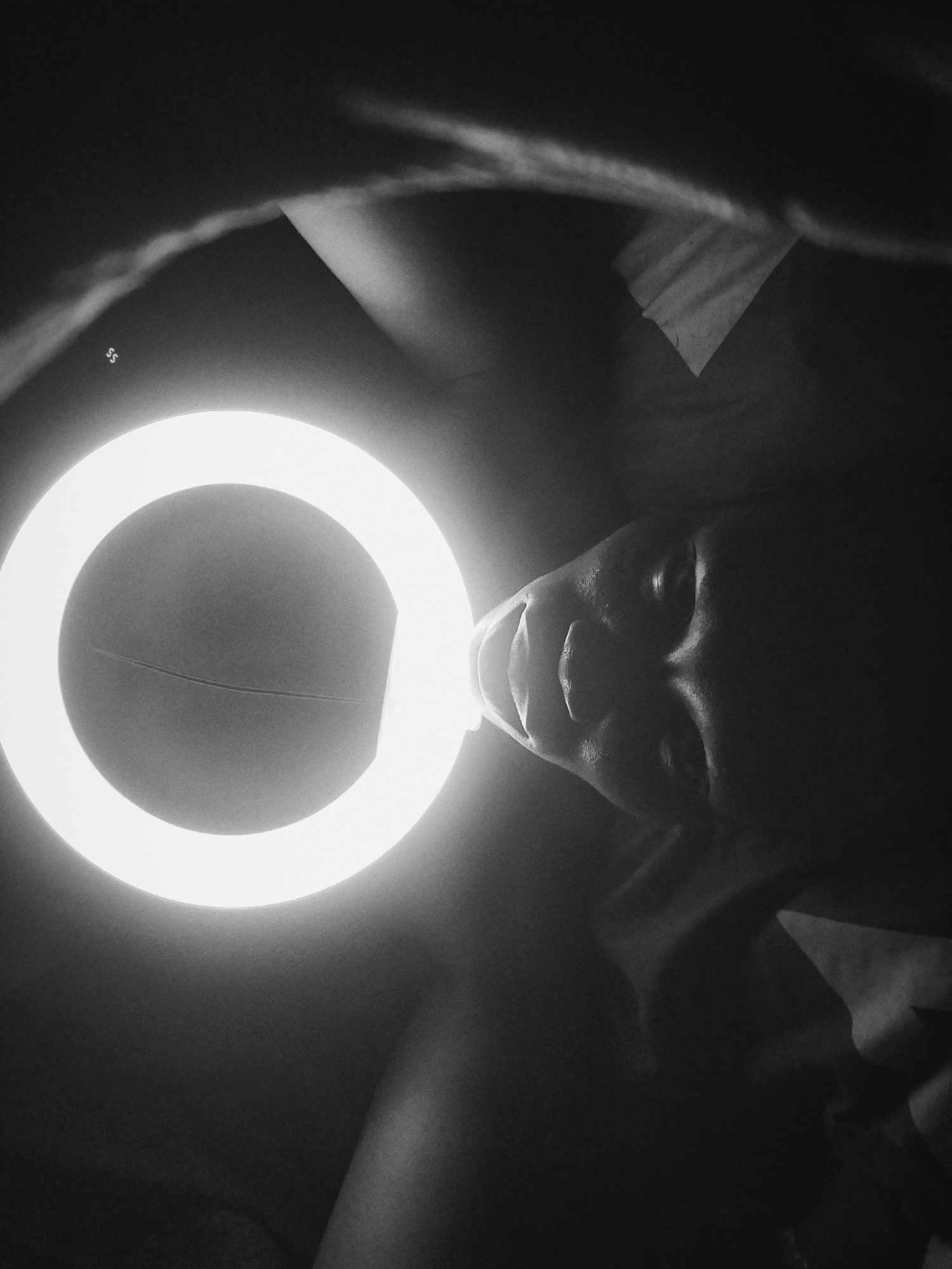 A black and white, low-angle photograph depicts a person's face illuminated by a ring light. The person is looking upwards, with the bright light positioned directly in front of their face, casting strong shadows. The background is dark and indistinct, revealing hints of fabric or bedding. An unclear mark or symbol is located on the top left of the ring light. The location and city are unknown.