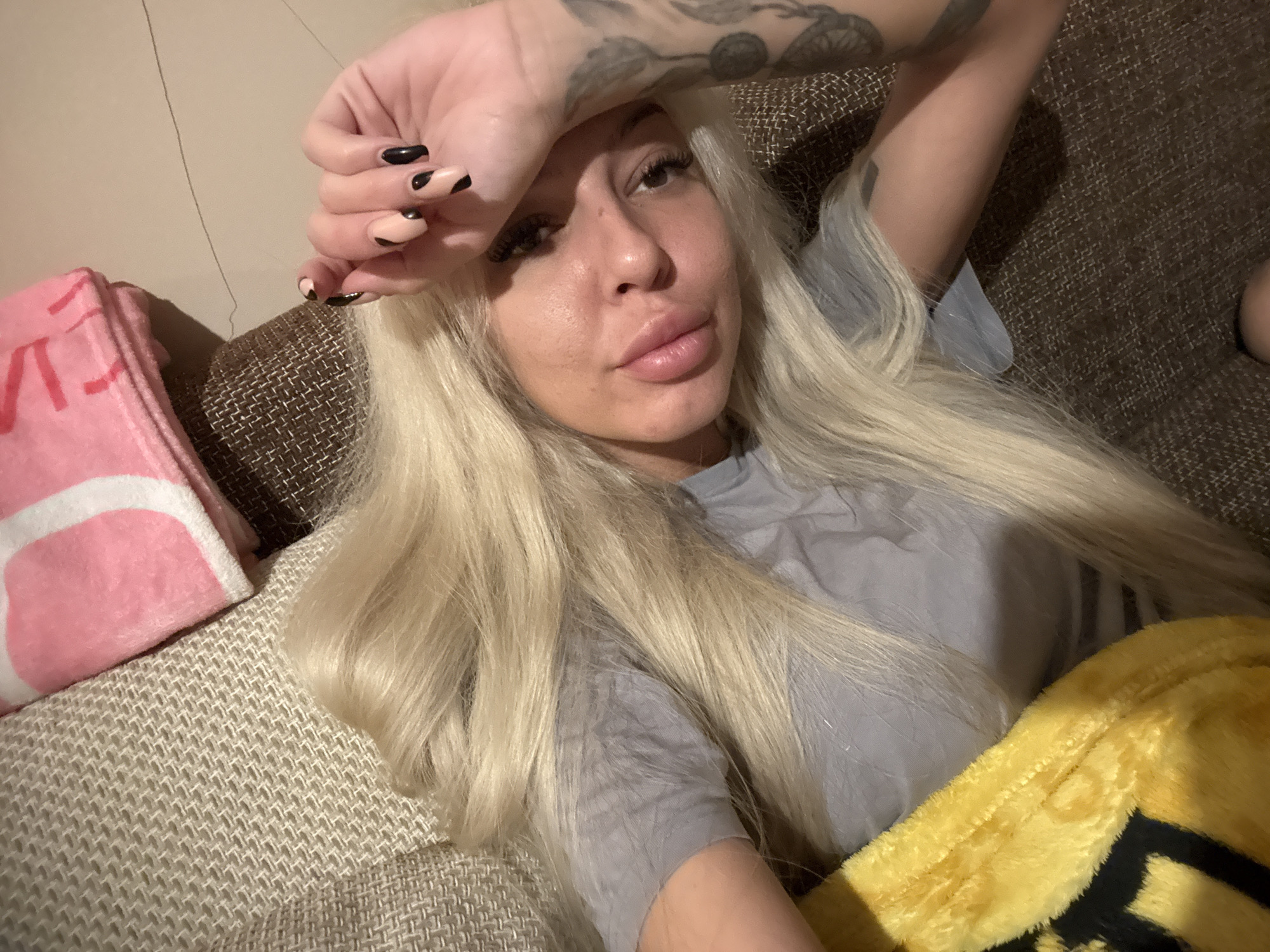 A woman with long blonde hair is lying on a sofa. She has tattoos on her arm and black nail polish. She is wearing a gray shirt. A pink and white blanket is on the left side of the sofa and a yellow blanket is on the right. The location is unknown.