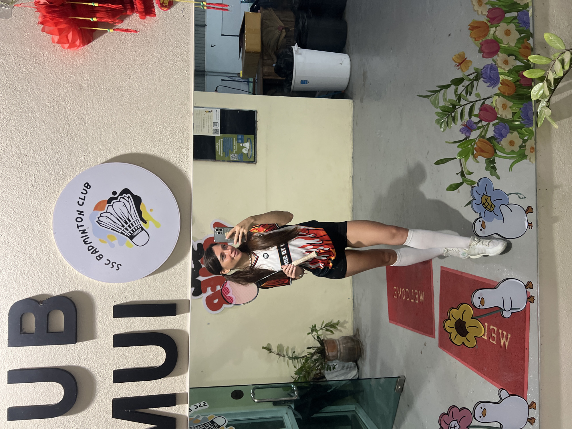 A woman is taking a selfie in a mirror. She is wearing a black and orange shirt, black shorts, and white knee-high socks. She is holding a smartphone in her right hand and a badminton racket in her left. The mirror is in a room with a sign that says "SSC BADMINTON CLUB." There is also a potted plant in the room and a red welcome mat with a flower cartoon. The exact location is Unknown, Unknown.