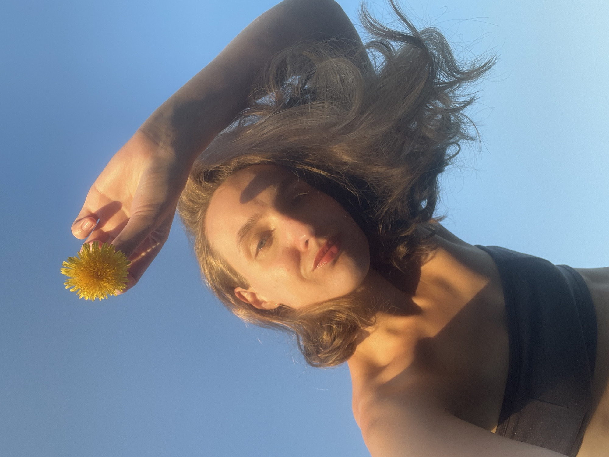 A low-angle shot depicts a woman holding a yellow dandelion flower against a clear blue sky. The woman has fair skin, shoulder-length brown hair, and is wearing a black top. The light source appears to be sunlight, casting shadows on her face and body. The image was captured outdoors, with the sky as the background. There is no verifiable evidence to support that this photograph was taken in the city of Подшивалово, Russia.