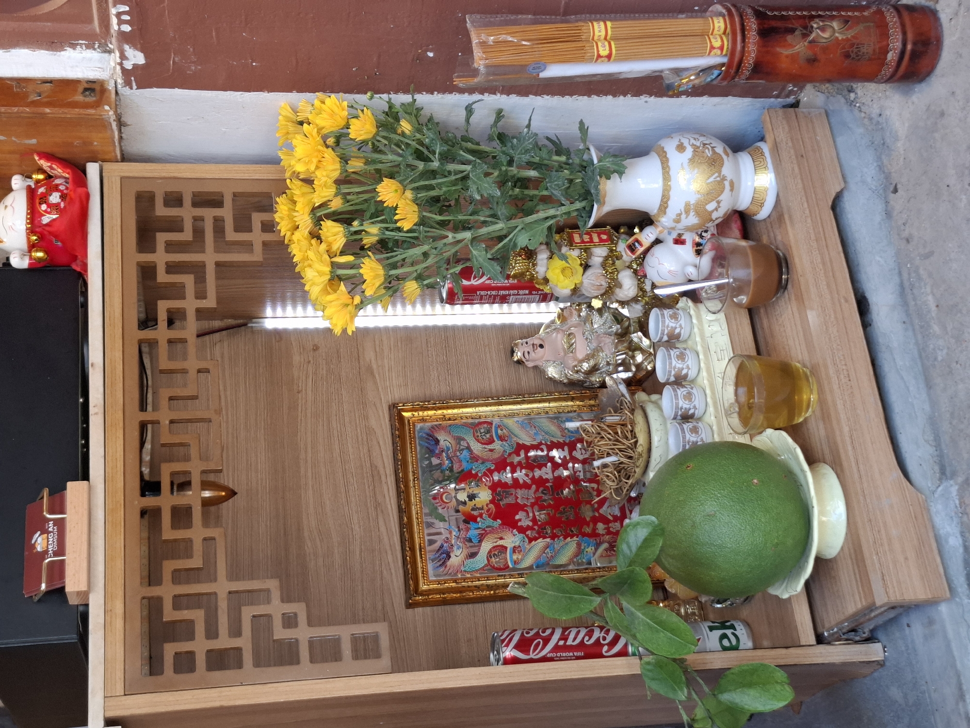 The image depicts a small altar with various religious and decorative items. The altar is made of wood and has intricate carvings on its upper section.
On the altar, there is a framed picture with a red background and intricate gold and blue dragon designs. To the left of the framed picture is a can of Coca-Cola. In front of the framed picture, there is a small brass bowl containing what appears to be incense sticks or dried herbs.
To the right of the framed picture, there is a statue of a seated figure in golden robes, possibly a deity or a Buddha statue. Next to this statue, there is a decorative arrangement of what seems to be white and gold baubles, topped with a yellow flower. A small white vase with golden dragon motifs holds a bouquet of bright yellow chrysanthemums.
In the foreground, a large green pomelo rests on a decorative white dish. To the right of the pomelo, there is a small glass of amber-colored liquid, likely tea. Further to the right, a dark glass contains a brown liquid, possibly coffee, with a small spoon in it. Behind these, there are several small white cups arranged in a row.
On the far right of the image, a cylindrical container holds a bundle of yellow incense sticks.
The altar is placed on a wooden shelf structure. The background suggests an outdoor or semi-outdoor setting with a weathered wall to the right. The floor is concrete.
The image is taken from a medium-low angle, focusing on the altar and its contents. The lighting suggests daylight. There are no people visible in the image. The overall scene appears to be a place of worship or a personal shrine.
While the image provides a detailed view of the altar, there are no explicit visual cues that definitively place it in Nha Trang, Vietnam, or any other specific city. However, the presence of items like incense, pomelo, and the style of the altar could be commonly found in Southeast Asian countries, including Vietnam.