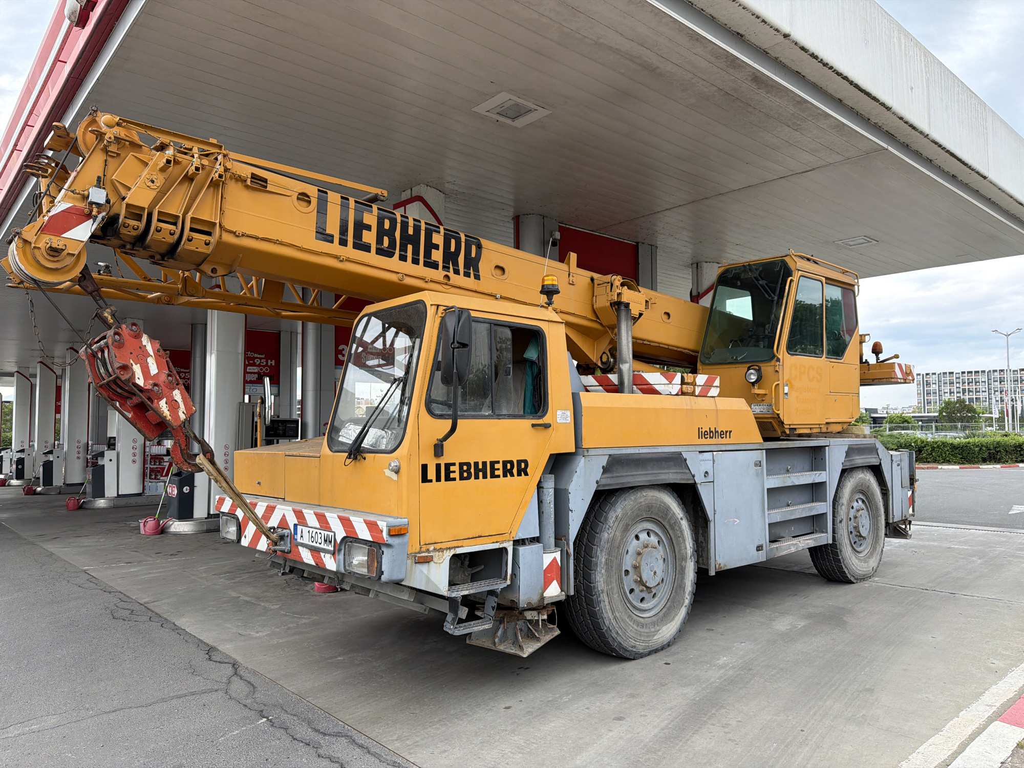 The image shows a yellow Liebherr crane truck parked under the canopy of a gas station. The crane arm is extended over the front of the vehicle, and the hook is visible hanging down. The truck's license plate reads "A 1603 MM." The backdrop includes gas pumps, a building, and a street with some greenery in the distance. The text "Diesel 1-95H" and the number "3" are visible on the gas station. The environment suggests the location is a gas station forecourt in the city of Burgas, Bulgaria.