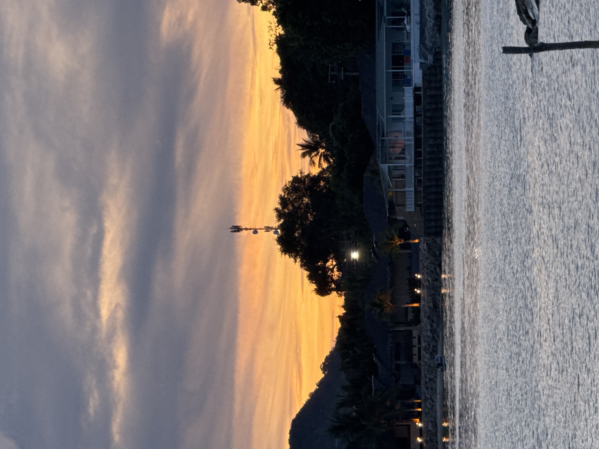 The image depicts a coastal scene in Baan Tai, Thailand, captured during sunset. The sky transitions from gray to orange, with clouds dispersed throughout. A cell tower is visible in the distance. The foreground features the shoreline, lined with trees, buildings, and illuminated pathway lights. Water occupies a significant portion of the right side of the image.
