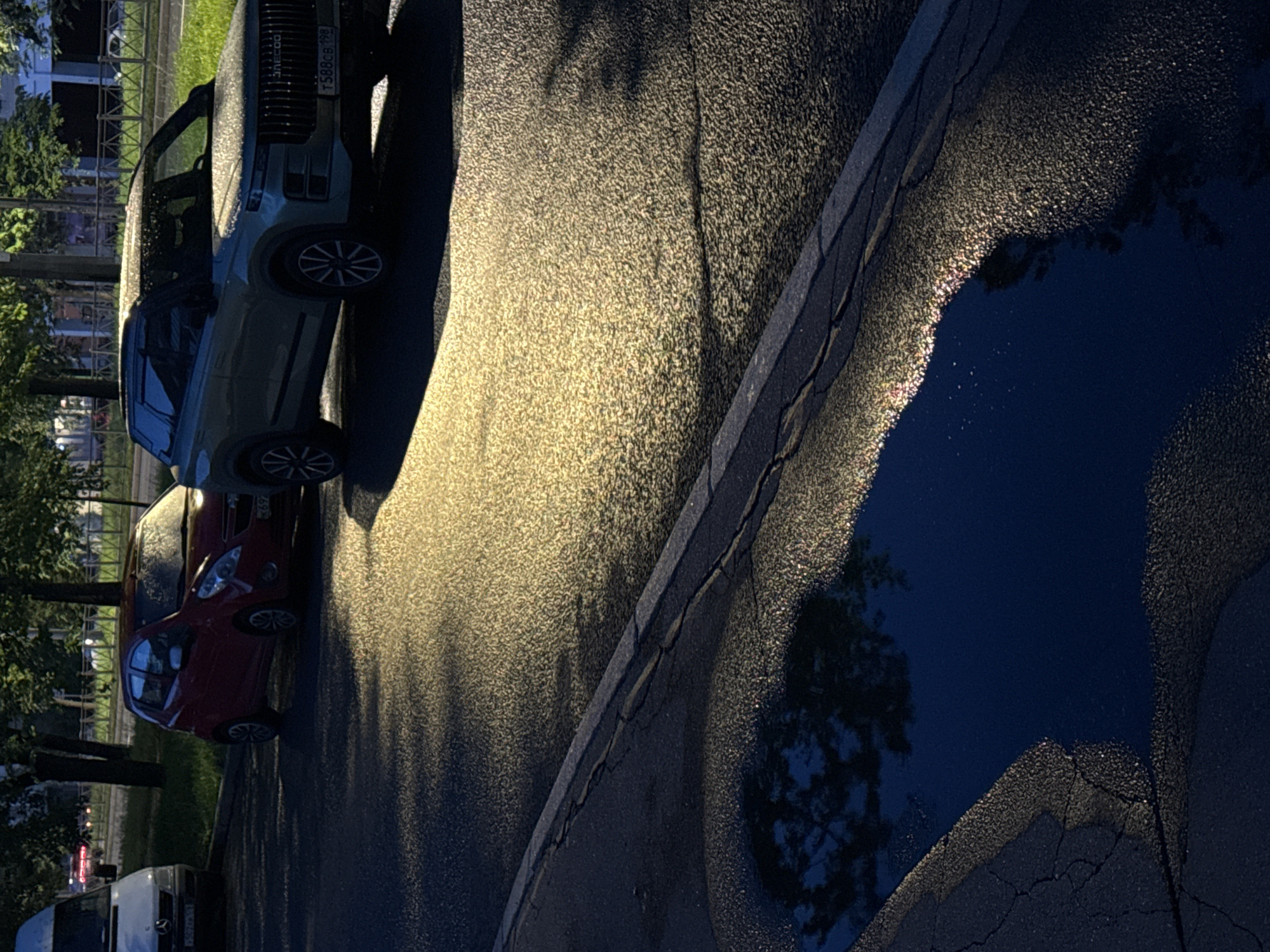 The image depicts a street scene in Saint Petersburg, Russia, likely during daylight hours with a reflective wet surface, possibly due to recent rain. In the foreground, a dark puddle with a sharp edge reflects the sky and trees, with iridescent highlights on its surface. The asphalt road exhibits cracks and a textured surface, illuminated by a strong, angled light source casting shadows.
Parked on the right side of the road is a modern, light-colored SUV with a distinctive grille and license plate T588CB 198. The SUV's roof and hood are speckled with water droplets. To the left of the SUV is a small, red hatchback car, also parked. Further to the left, a white van is partially visible. Trees line the background, casting shadows on the road and grass. The overall atmosphere is calm and quiet, with no visible people or ongoing activities. The lighting suggests either early morning or late afternoon sun.
