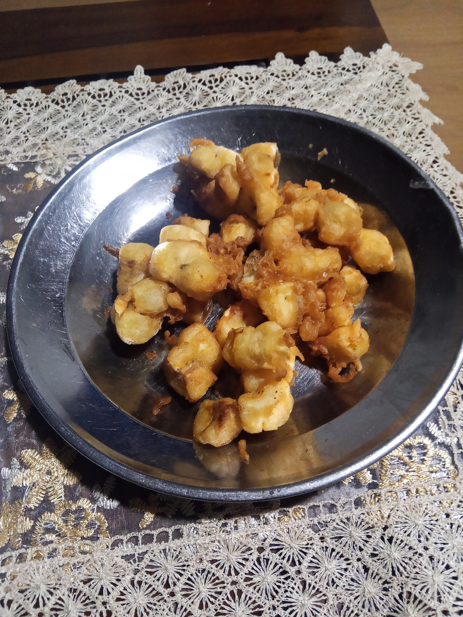 A silver metal plate filled with golden-brown fried food is centrally placed on a table. The food appears to be bite-sized pieces, possibly battered and deep-fried. The plate is set on a decorative lace tablecloth with intricate patterns in white and gold, which is laid over a darker surface. In the background, a wooden surface is visible. The lighting suggests an indoor setting, likely a dining area. The image does not provide any specific indicators about the city of Makale, Indonesia.