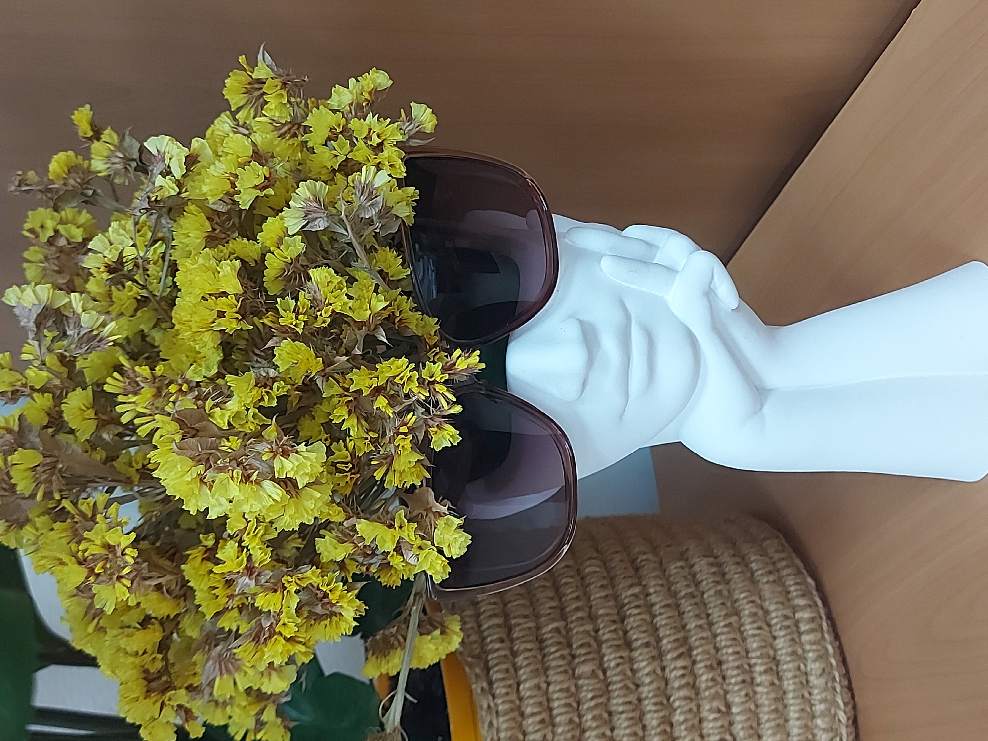 The image depicts a white plaster bust wearing sunglasses, with a bouquet of yellow flowers arranged around it. The bust and flowers are positioned on a brown woven basket, resting on a light brown wooden surface. The background is a similar light brown wood. The scene is well-lit and appears indoors. There is no indication that the photograph was taken in Chaikovsky, Russia.