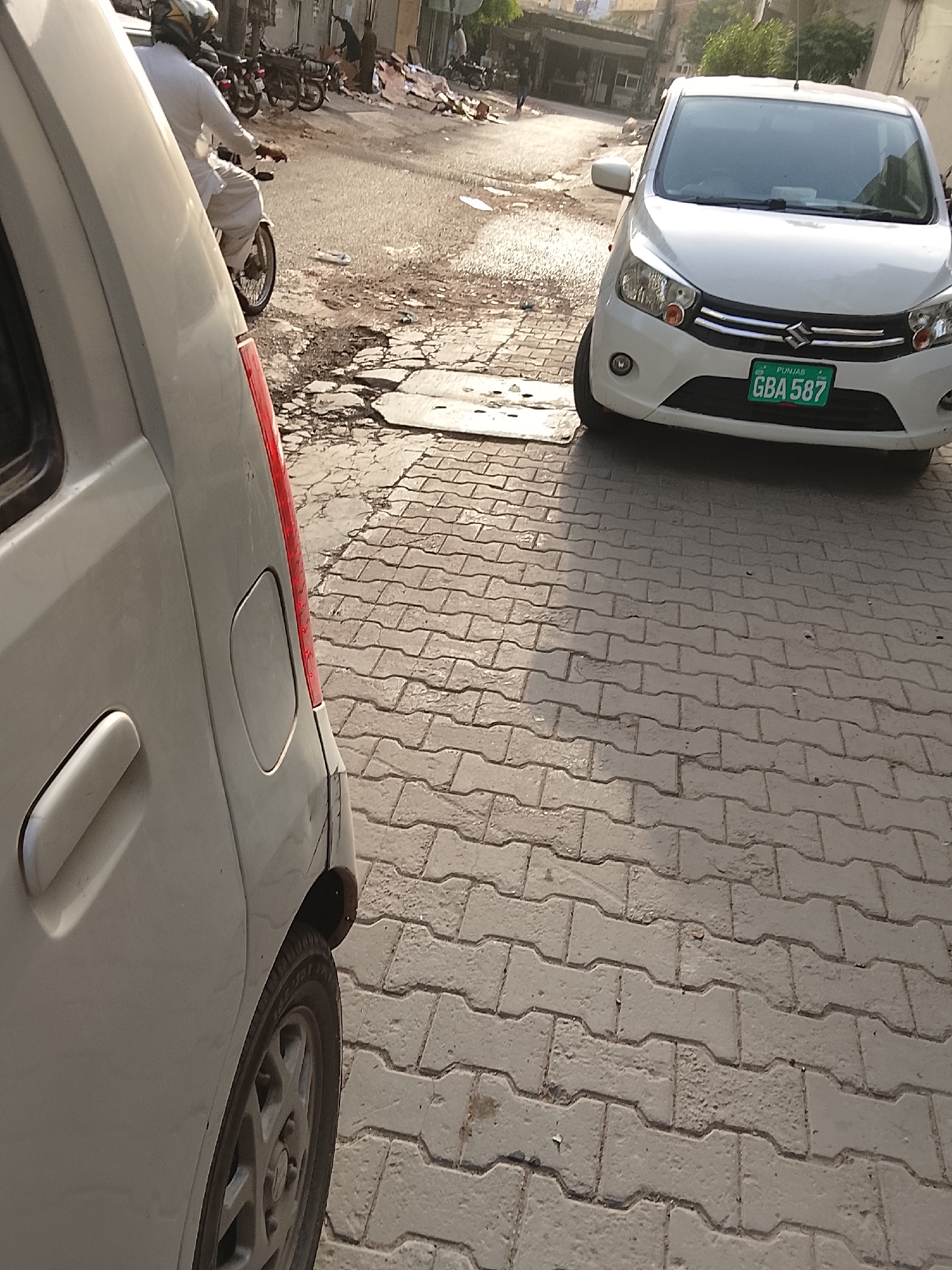 The image shows a street scene in Gulgasht Colony, Pakistan. In the foreground, the rear portion of a white car, including its taillight, door handle, and rear tire, is visible. Adjacent to it, further into the frame, a white Suzuki Cultus car is parked. The car has a green license plate with "GBA 587" written on it, and the word "Punjab" is visible on the top of the license plate. In the background, a motorcyclist in white clothing and a helmet is driving on the street. Several parked motorcycles are visible. Debris is visible on the road near the motorcycles. In the distance, there are buildings and a few people are standing. The street is paved with interlocking pavers, and the road has some patches of damage.