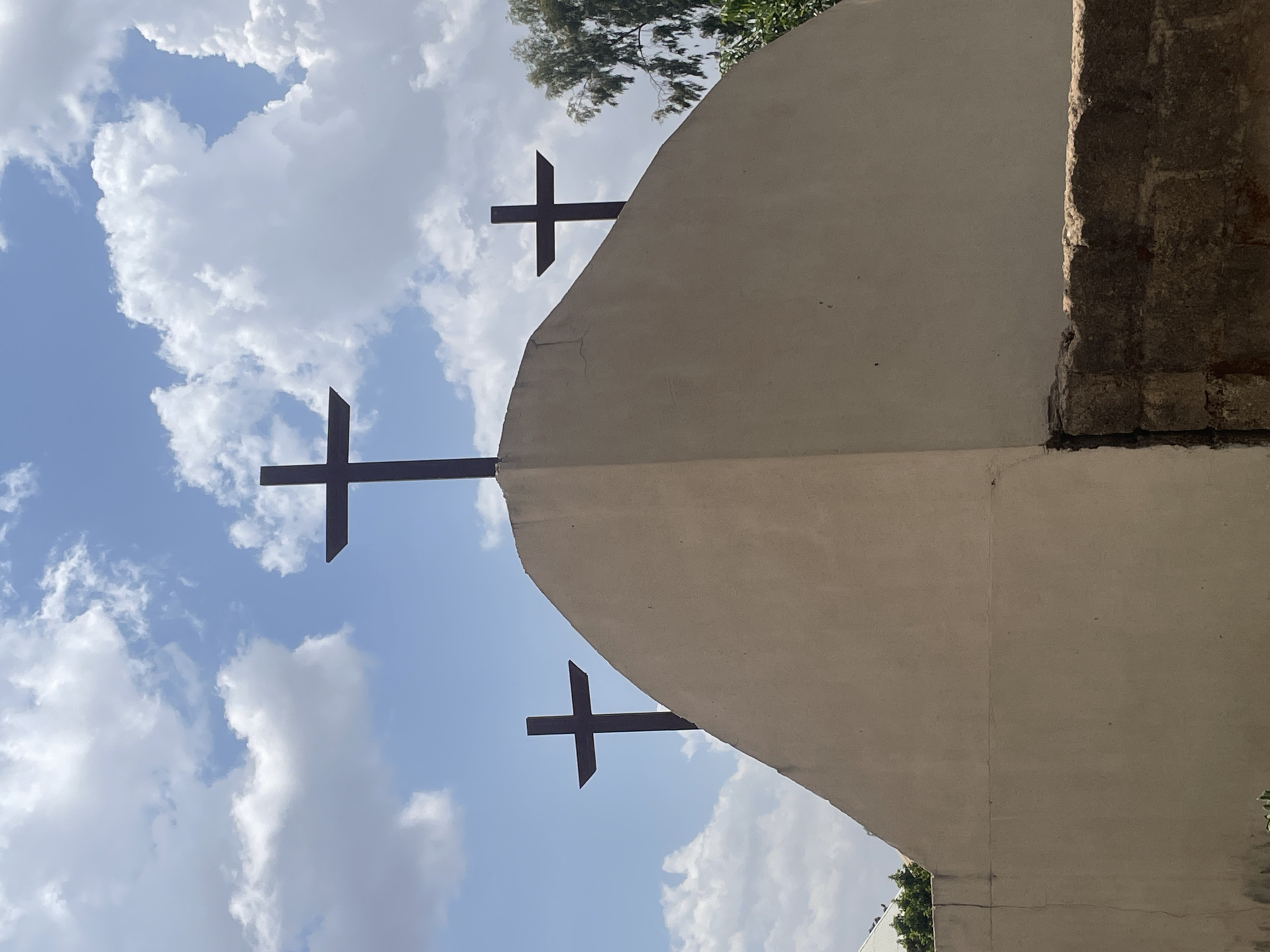 The image shows an upward view of a building structure in Jos, Nigeria, with a light-colored, possibly beige or white, concrete or stucco facade. Three dark, metal crosses are affixed to the facade, spaced vertically along its curvature. A segment of a rough, stone or brick wall is visible on the right edge of the frame. The background is a blue sky with scattered white clouds. Green foliage and branches are visible near the top of the building.