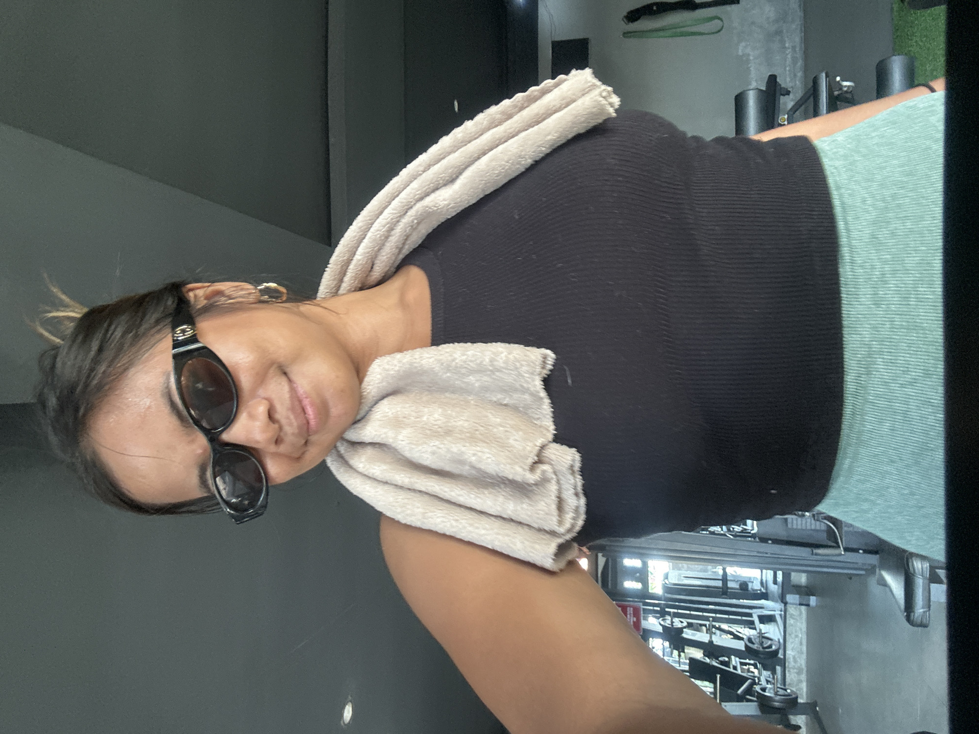 A woman with dark hair pulled up in a bun and wearing black oval sunglasses and a black ribbed tank top poses for a selfie in a gym. A light beige towel is draped around her neck. She is also wearing light green ribbed athletic bottoms. The background shows gym equipment, including weight racks and benches, and appears to be in an indoor setting. The overall lighting suggests it could be daytime. The woman is smiling and looking at the camera.