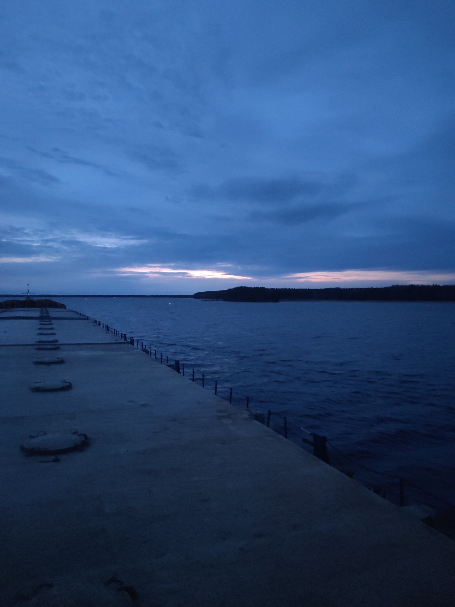 The photograph depicts a pier or breakwater extending into a body of water, possibly a lake or river. The time of day appears to be dusk or early evening, given the low light and blue-toned sky. The sky exhibits a gradient of color, with a lighter band near the horizon and darker hues above. Clouds are present, adding texture and depth to the sky. The body of water is dark and slightly choppy, reflecting the dim light. A strip of land with trees lines the horizon. The pier is made of concrete and has evenly spaced circular features, possibly manhole covers or access points. There is a railing along the pier's edge on one side.
While the provided location is Topornya, Russia, there is no explicit visual confirmation of this from the image itself.