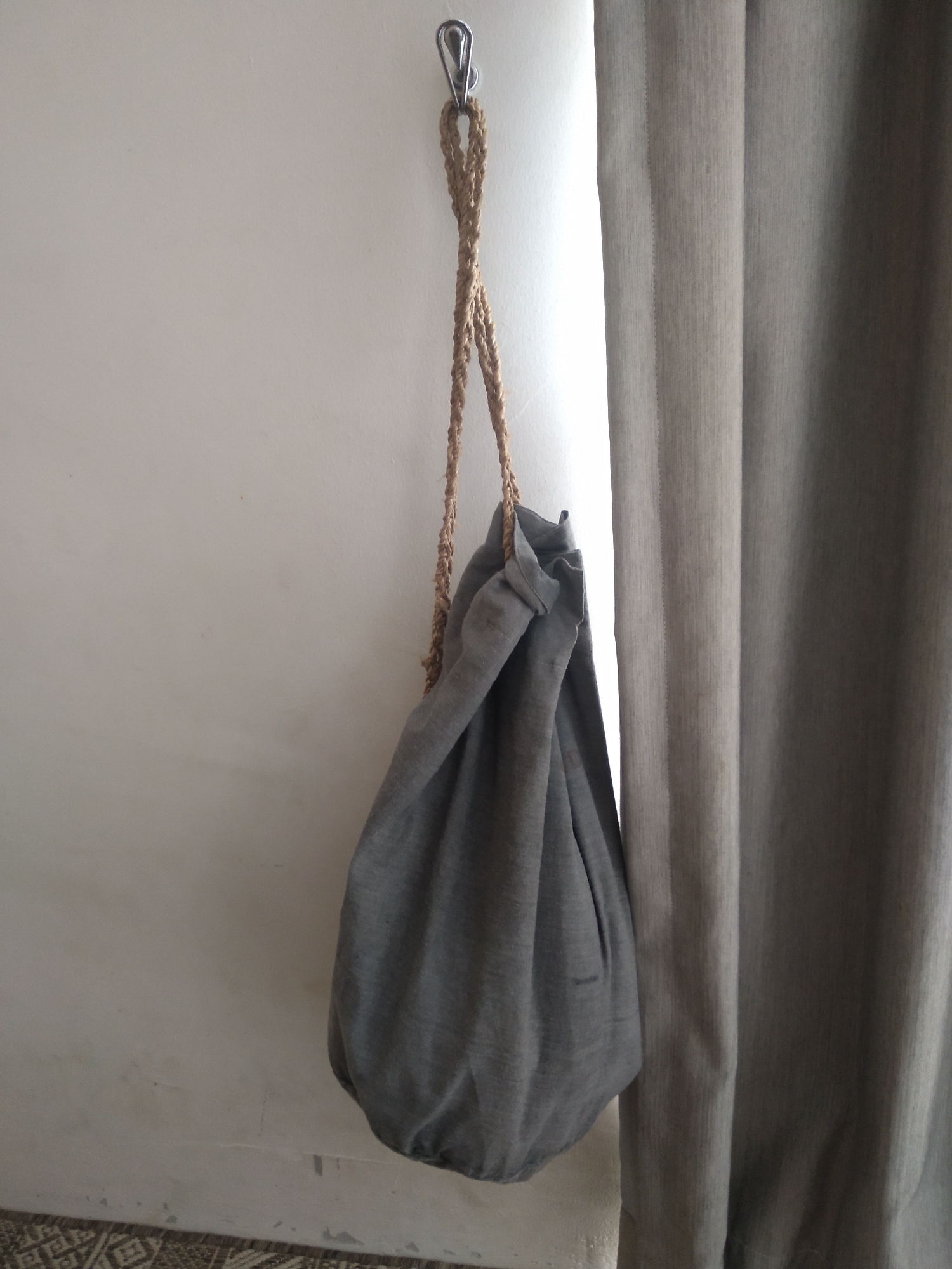 The image shows a grey drawstring bag hanging from a silver clip attached to a white wall. The bag is made of a textured fabric and has a braided rope handle. To the right of the bag is a grey curtain. The floor below the wall has a patterned rug. The scene is indoors, suggesting it could be a residential or commercial space. There is no indication of it being located in South Tangerang, Indonesia.