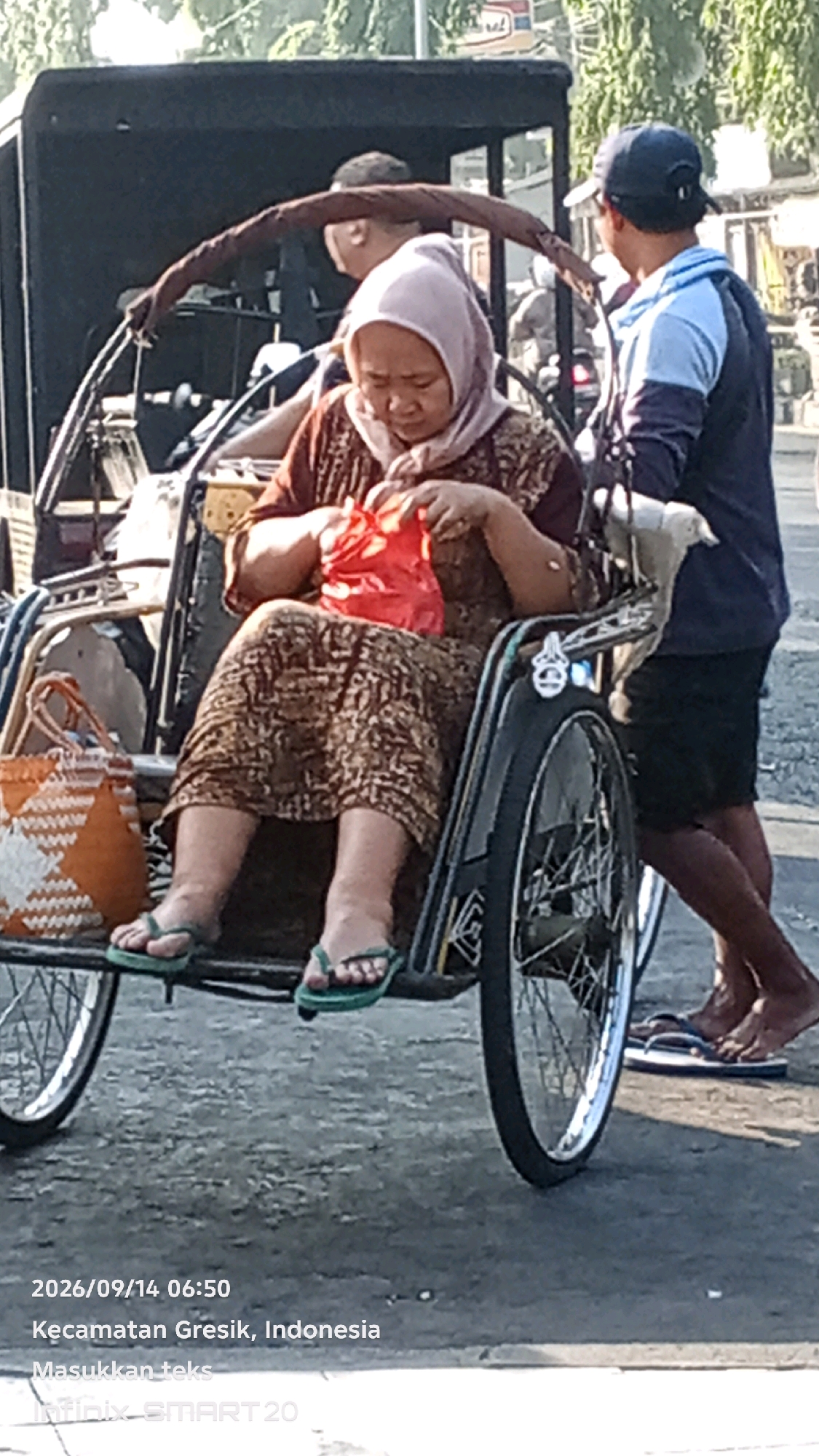 An outdoor, medium-shot captures a woman sitting in a pedicab, holding a red bag. The woman, wearing a light pink headscarf and a patterned brown dress, is seated and looking down at the bag. She has green flip-flops on. Next to her is an orange and white woven bag. A man in a blue shirt and black shorts is pushing the pedicab from behind. The background shows a street with trees and buildings. The bottom of the image includes text indicating the date and location: "2026/09/14 06:50, Kecamatan Gresik, Indonesia". The environment appears to be urban.