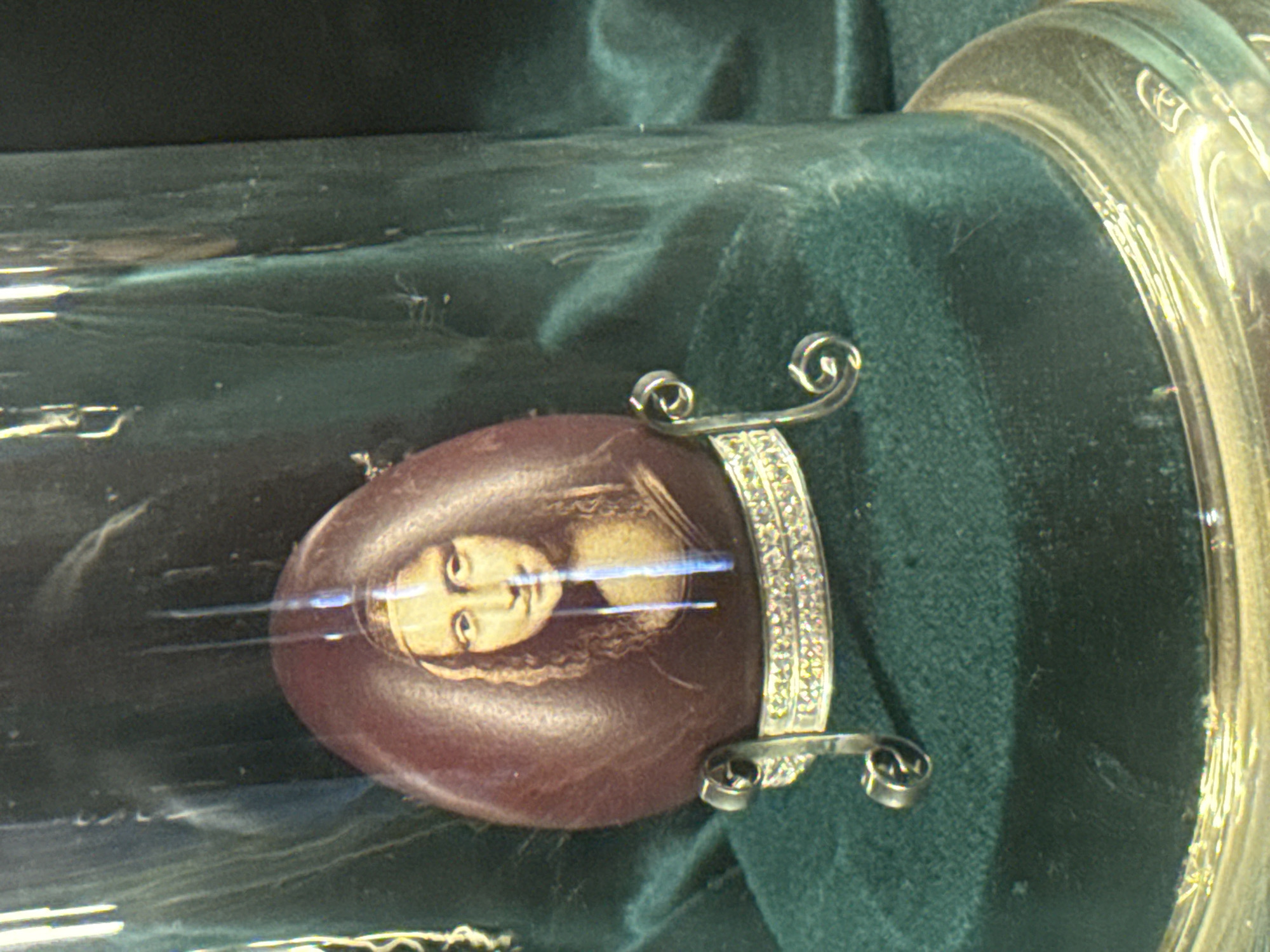 A close-up photograph shows an oval, maroon-colored object with an image of a person's face attached to a decorative, silver-colored metal setting. The object rests inside a clear, cylindrical container on top of a green, velvety fabric. The container appears to be housed within a museum or exhibit setting in the city of Chynadiiovo, Ukraine. The photograph focuses on the artifact itself, capturing the details of the image and its surrounding setting.