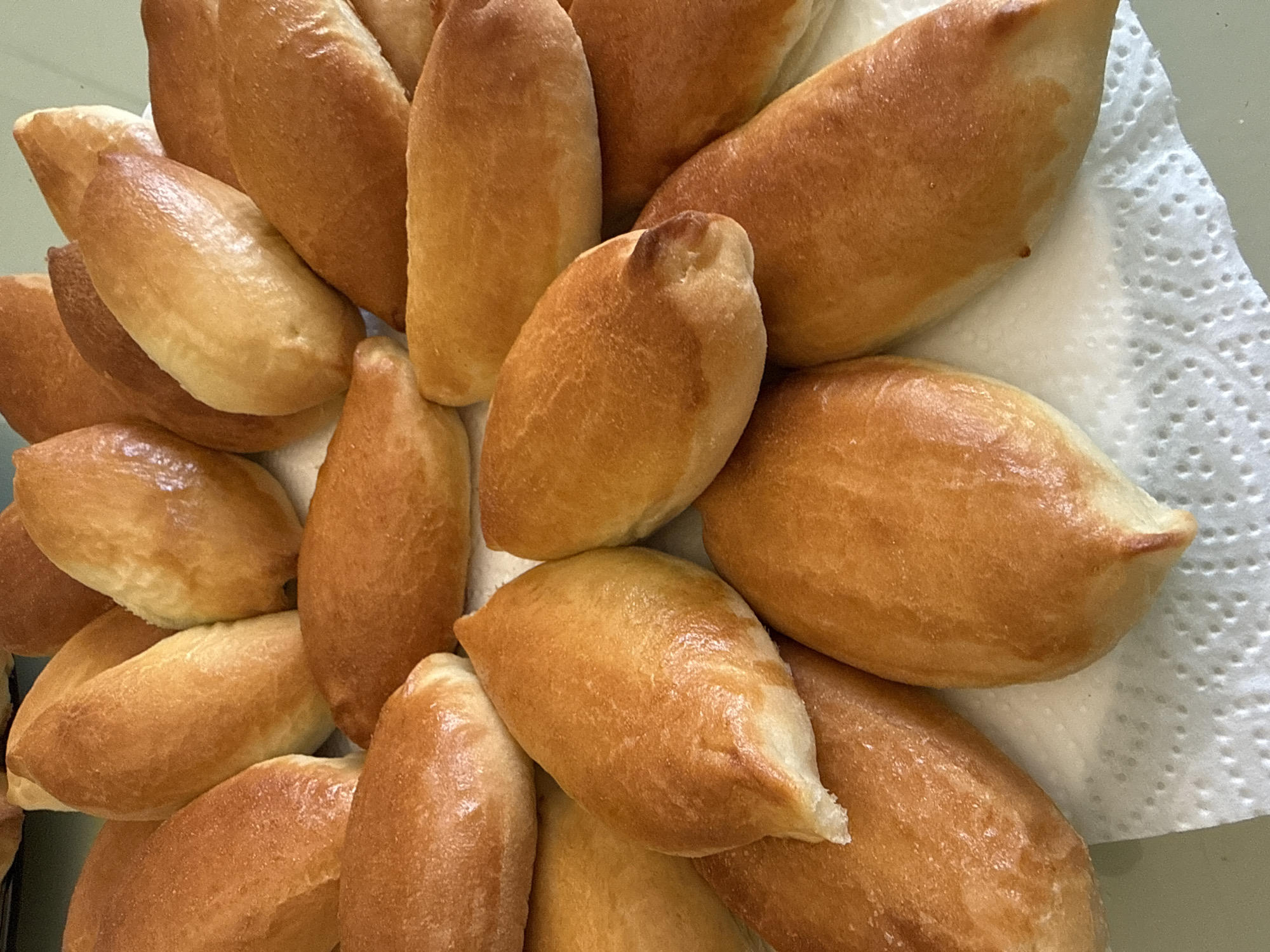 The image displays a close-up, top-down view of a large pile of freshly baked, golden-brown pastries. Each pastry is distinctively oval or spindle-shaped, tapering at both ends, and features a glossy, well-baked crust. The color of the crusts varies from a lighter golden hue to a deeper, rich brown, suggesting a thorough bake. The pastries are stacked somewhat haphazardly, filling most of the frame, indicating a generous batch has been prepared.
A white paper towel with a visible dotted texture is present underneath some of the pastries on the right side and bottom of the frame, suggesting they may be cooling or draining any residual oils. The appearance of the pastries evokes a sense of warmth and freshness, commonly associated with recently prepared baked goods. Given the location context of Votkinsk, Russia, these pastries are likely *pirozhki*, a popular traditional Russian dish. There are no human subjects, activity, or visible text in the image.