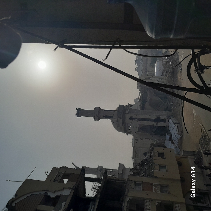 The image depicts a scene of destruction, likely from war or a major disaster. Buildings are severely damaged or completely destroyed, with rubble scattered across the landscape. A mosque or religious structure, though damaged, stands prominently amidst the wreckage. The scene is washed in a hazy, desaturated light, possibly from dust or smoke. A glimpse of sunlight penetrates the gloom. The foreground includes parts of a structure, likely a window frame, from which the photograph was taken.