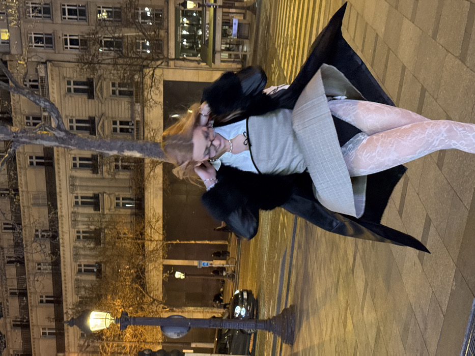 The image depicts a nocturnal street scene on a paved sidewalk in Paris, France, characterized by multi-story Haussmannian-style buildings illuminated in the background. A female subject with light-colored hair, wearing eyeglasses, a light-colored collared shirt, a grey strapless top with a black bow, a long black coat with faux fur collar and cuffs, a grey plaid skirt, black brief-style undergarments, and white lace-patterned tights, is captured in motion. Her skirt is visibly elevated by wind, exposing her undergarments and tights. Her hands are positioned near her head, indicating an action of adjustment or reaction. A traditional black lamppost with an illuminated globe is prominent on the left side of the foreground. A dark vehicle is partially visible on the street in the mid-ground, which features a white-striped pedestrian crossing. Deciduous trees with bare branches are present along the thoroughfare. Distant figures of pedestrians and a bicycle are discernible further down the street.