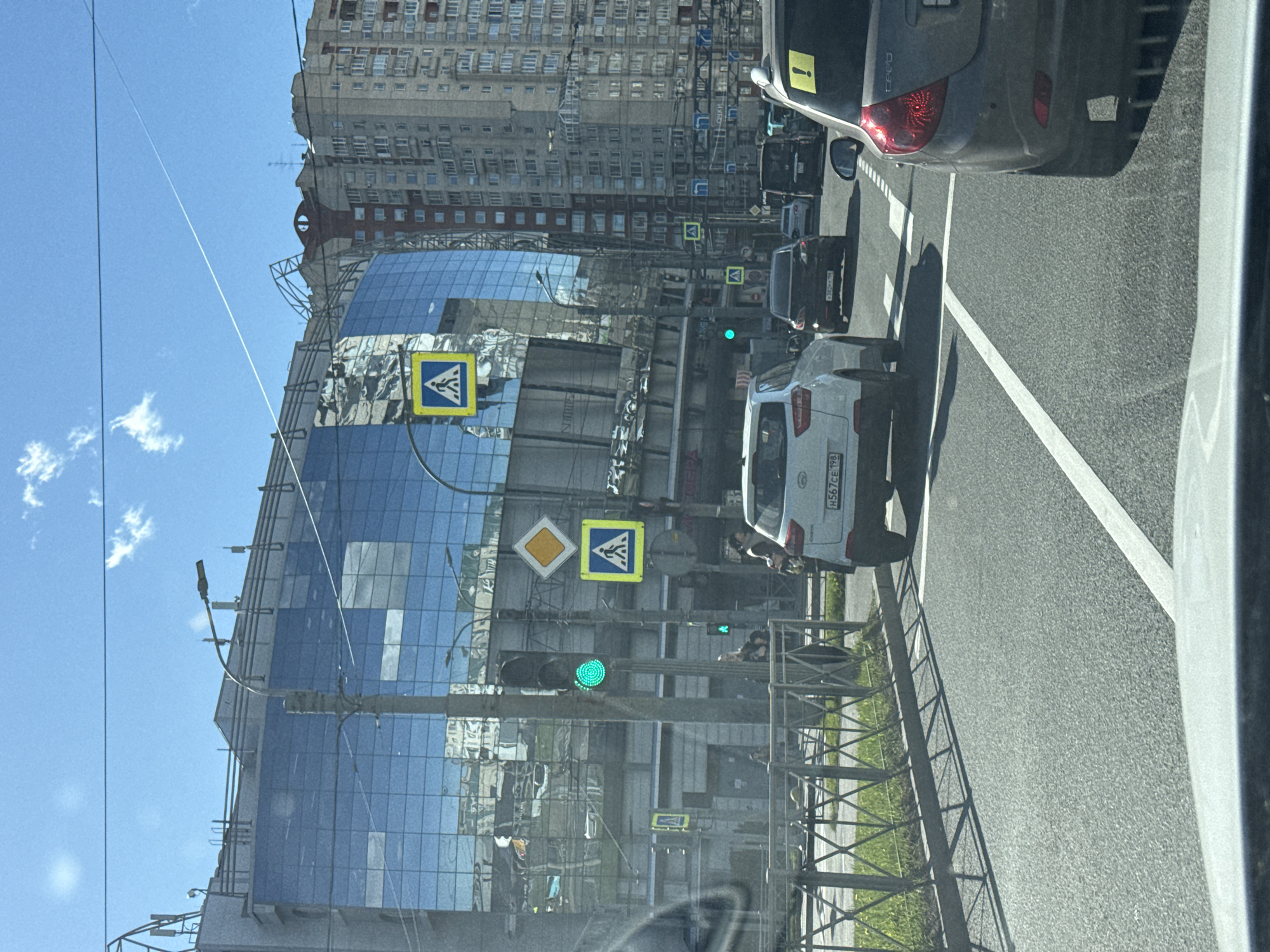 The image shows a street scene in Saint Petersburg, Russia. A modern glass building is on the left, reflecting the sky and surrounding buildings. Pedestrian crosswalk signs are visible near a green traffic light. Several cars are on the road, including a white car with the license plate "H567CE 198" and a gray Kia Ceed with a yellow "!". There are also other cars, like a black one, further down the road. A guardrail separates the road from a grassy area. The sky is clear and blue with some white clouds. Taller buildings are visible in the background.