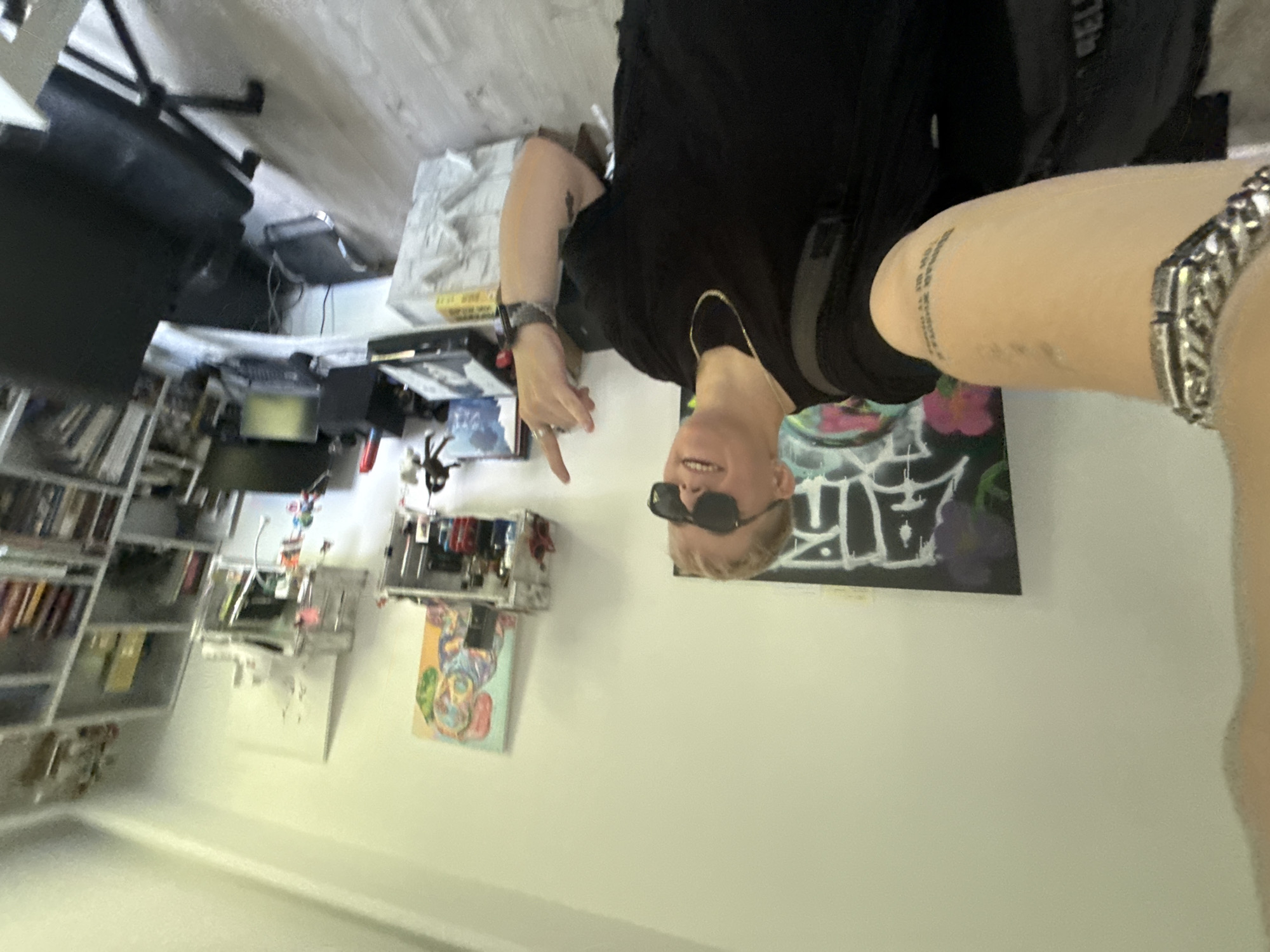 The scene is an indoor room located in Sofia, Bulgaria. A light-skinned individual with short blonde hair and black sunglasses occupies the center-left foreground, likely captured in a selfie. The person is wearing a black t-shirt, a gold chain necklace, a silver link bracelet on the left wrist, and a dark-colored watch on the right wrist. Tattoos are visible on both forearms, including discernible text on the left forearm. A black backpack is worn. The individual is smiling and raising their right index finger upwards.
The background features white walls and a light-colored wooden or laminate floor. On the left wall, a large black canvas displays white graffiti-style lettering and pink/purple accents. To the right, a colorful, abstract painting of an animal (possibly a dog) is mounted above a small, open white shelf unit containing various bottles and small objects, including a red figurine. Further right, a tall white bookshelf filled with books and miscellaneous items stands against the wall.
A desk area is visible to the right, featuring a white desk with a laptop, an external monitor, and various computer peripherals including speakers and cables. A black office chair with wheels is positioned in front of the desk. A metallic footrest rests on the floor beneath the desk. A white, possibly wrapped or boxed item is present on the floor to the right of the person's leg. The room is well-lit.