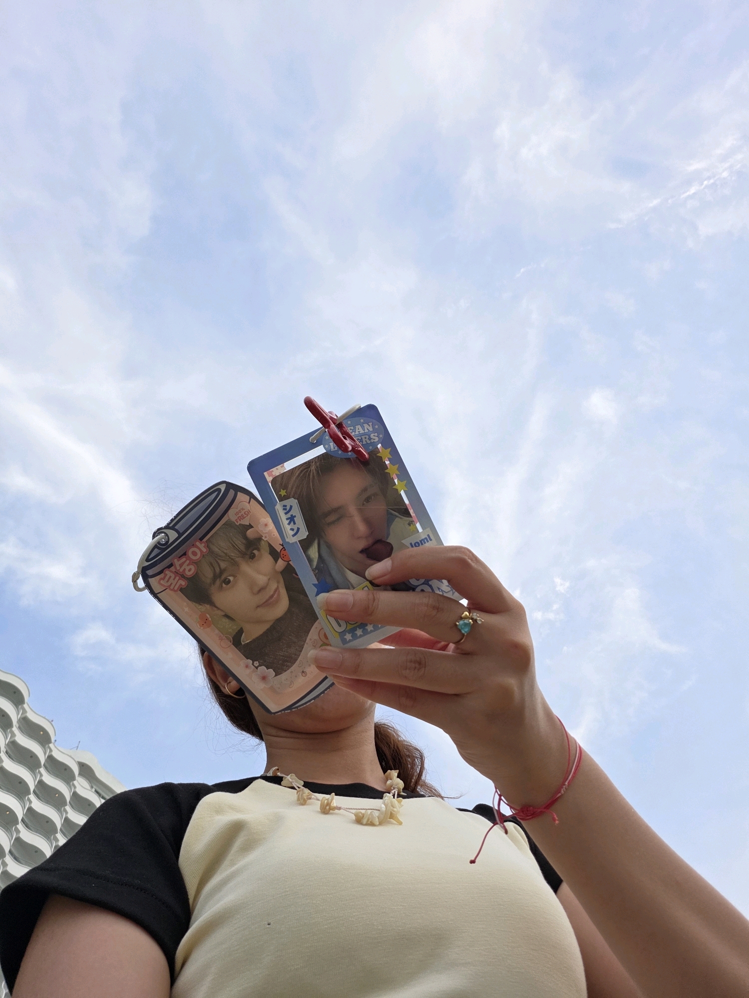 In George Town, Malaysia, a person holds up two K-pop photocards against a sky filled with white, wispy clouds. The individual is wearing a cream-colored shirt with black sleeves and a shell necklace. A red clothespin attaches the two photocards together. The photocards feature images of two young men, likely K-pop idols. The person is also wearing a ring with a blue stone and a red string bracelet on their right hand. In the background, the edge of a building with a white, wavy facade is visible.