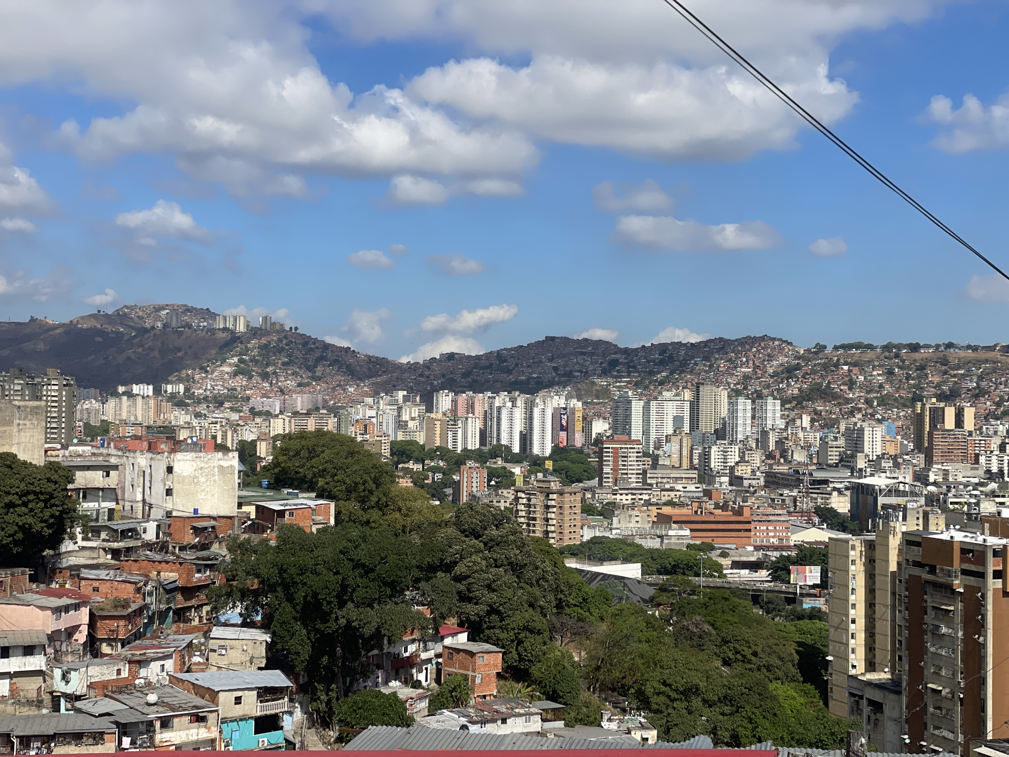 The image shows an elevated view of Caracas, Venezuela, showcasing a complex urban landscape. In the foreground, there are informal settlements characterized by closely packed, low-rise buildings with a mix of construction materials like brick and corrugated metal roofs. These structures appear to be built on a hillside.
Moving towards the middle ground, the city transitions into a more organized layout with mid-rise to high-rise buildings, typical of a more developed urban center. There are residential and commercial buildings with a variety of architectural styles.
In the background, the topography rises again, featuring more informal settlements clinging to the hillsides, interspersed with some taller buildings. The sky is blue with scattered white clouds. A cable runs diagonally across the upper right portion of the frame, suggesting the presence of a cable car system, a common feature in Caracas to navigate the hilly terrain. There are no people visible. The environment is densely populated with a mixture of poverty and urban density.
