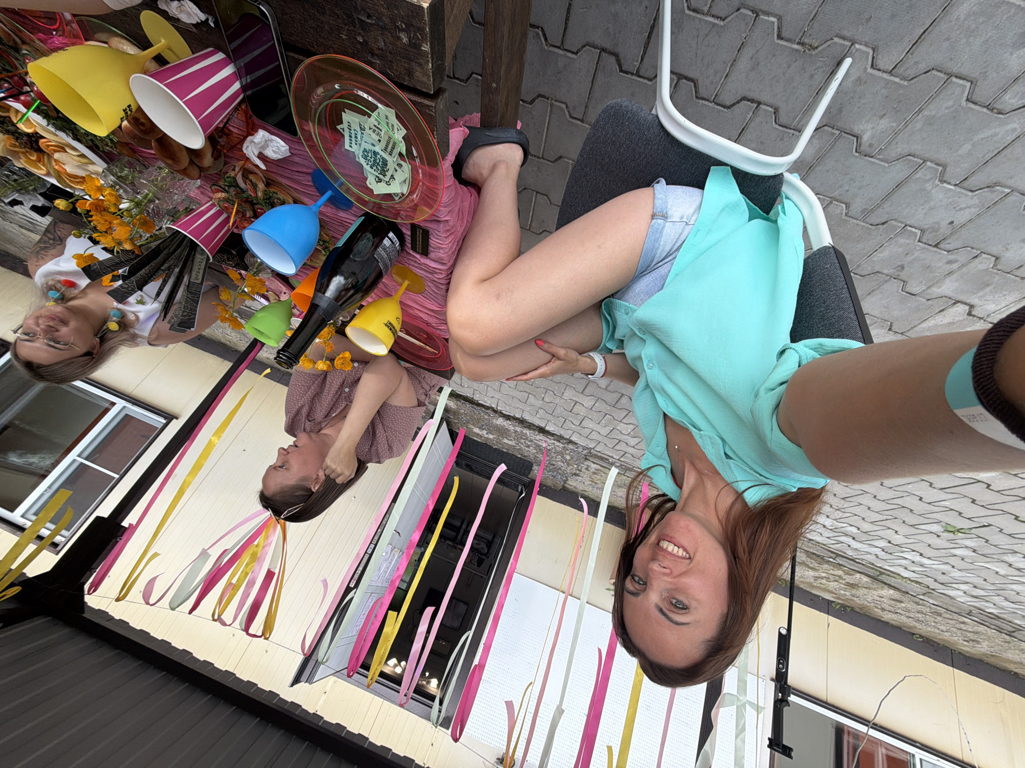 Two women are visible in this outdoor, daytime photograph, likely taken in Birobidzhan, Russia. The scene appears to be a casual gathering or party. In the foreground, one woman is seated, wearing a light blue top and denim shorts, and smiling at the camera. Her right hand is resting on her leg. Behind her, to the left, another woman wearing a pink polka-dot shirt is partially visible, her head turned away from the camera. A table is laden with various objects, including colorful cups (yellow, pink, blue, and green), a bottle of wine, a pink plate with what appears to be money or tickets, a bouquet of yellow flowers, and some food items. Colorful ribbons are also present, draped and hanging. The ground is paved with interlocking bricks. The overall mood seems festive and relaxed.