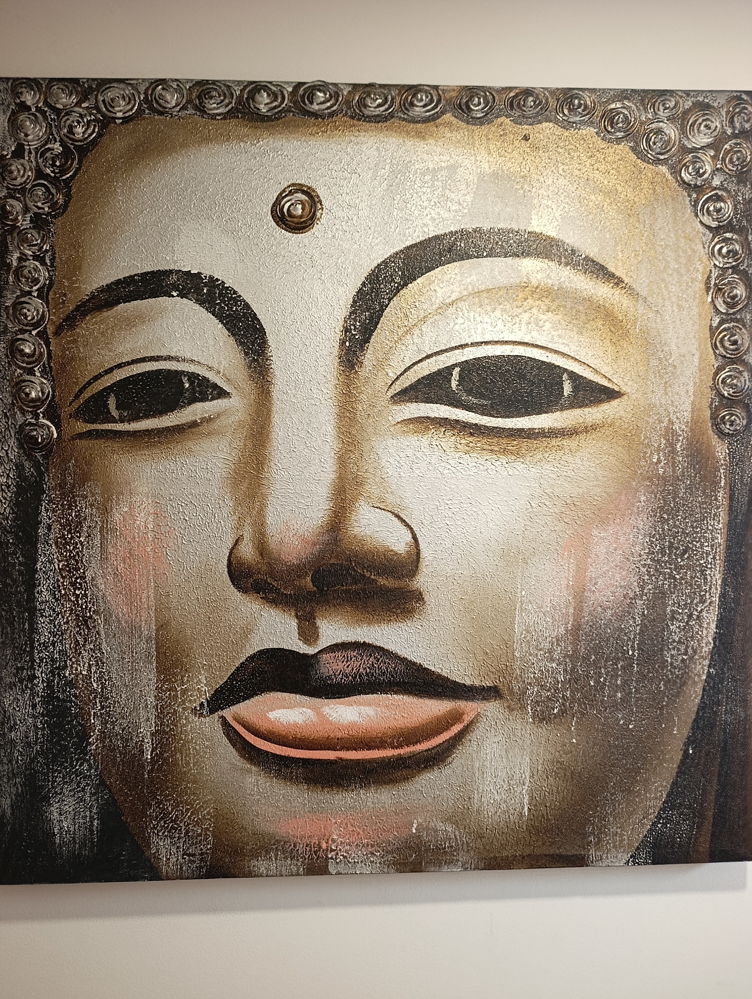 The image is a close-up, head-on painting of a Buddha's face. The painting features textured brushstrokes and uses a palette of gold, silver, white, black, and pink. The Buddha's eyes are closed, and a subtle smile plays on its lips. A circular mark, possibly representing the urna, is visible on the forehead. The painting has a decorative border of coiled motifs around the top and sides. The painting is displayed on a plain white wall, suggesting it is likely in a home or gallery setting. There is no indication of its location in the city of Setúbal, Portugal.